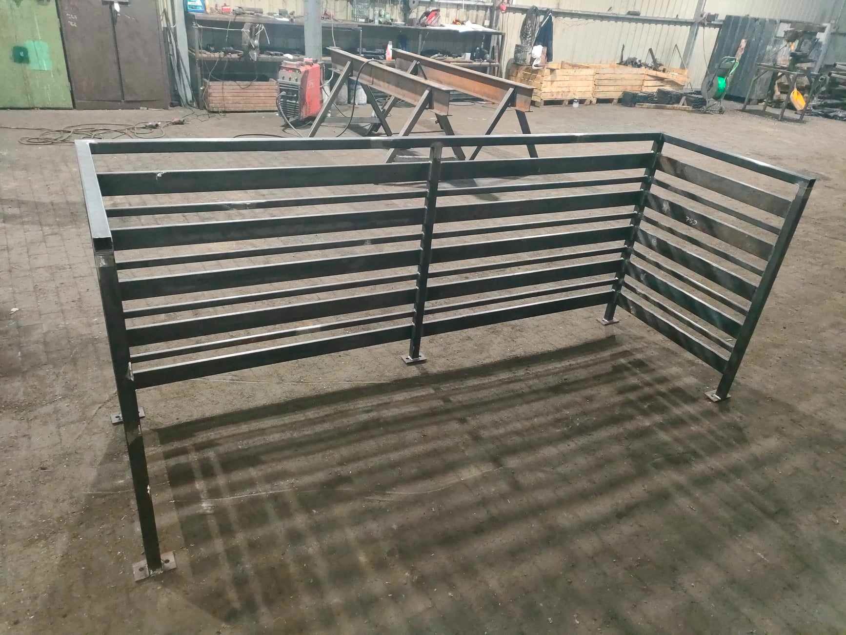 Surowa, metalowa konstrukcja ogrodzenia panelowego z poziomych profili, spawana w warsztacie, widoczna spawarka w tle.