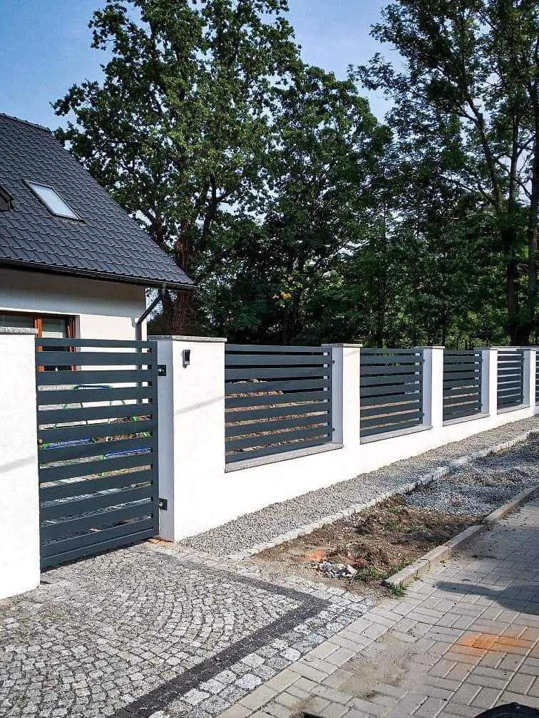 Nowoczesne ogrodzenie panelowe z szarej stali z bramą, osadzone na białym murku, widok z zewnątrz na posesję z kostką brukową i fragmentem chodnika.
