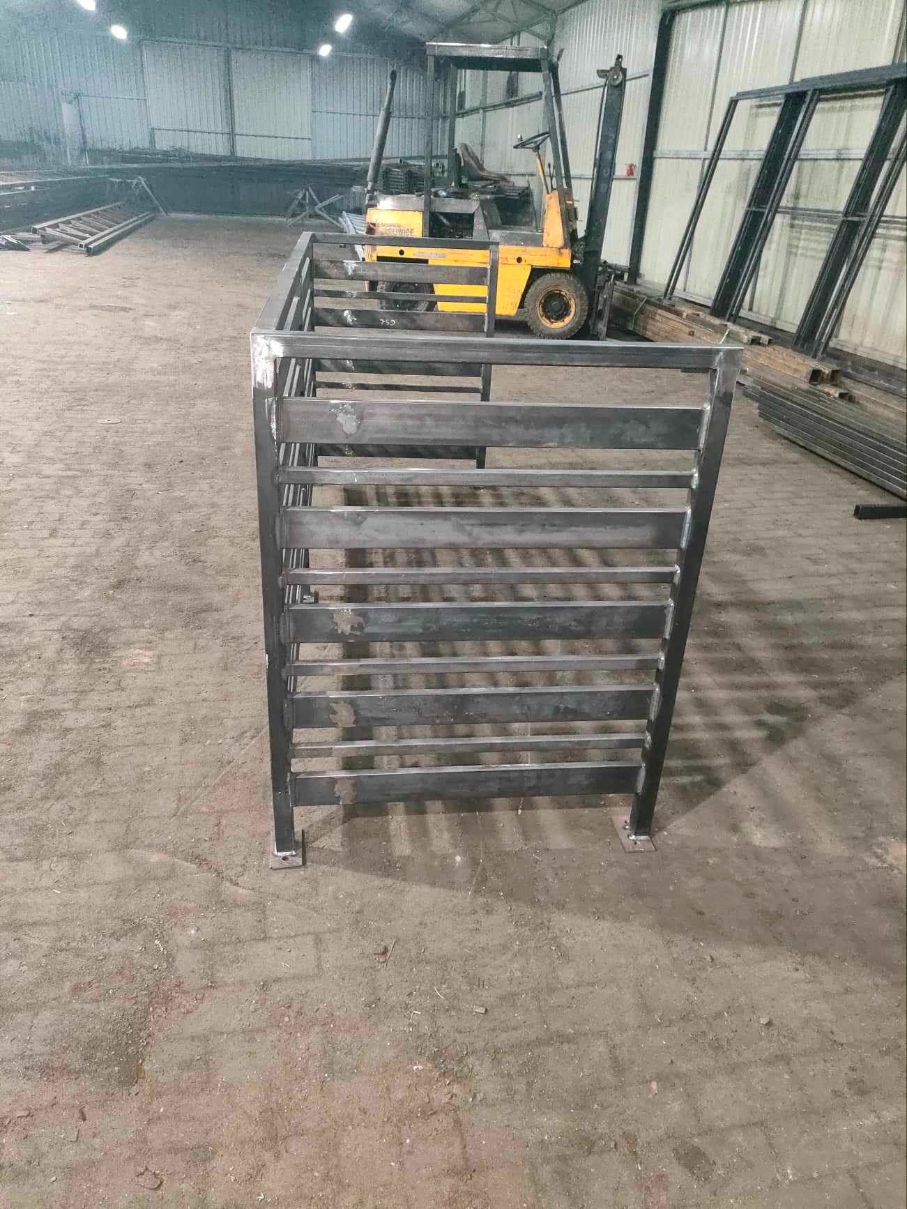 Surowa, metalowa konstrukcja klatki schodowej z poziomymi szczeblami, spawana w hali produkcyjnej, z wózkiem widłowym w tle.