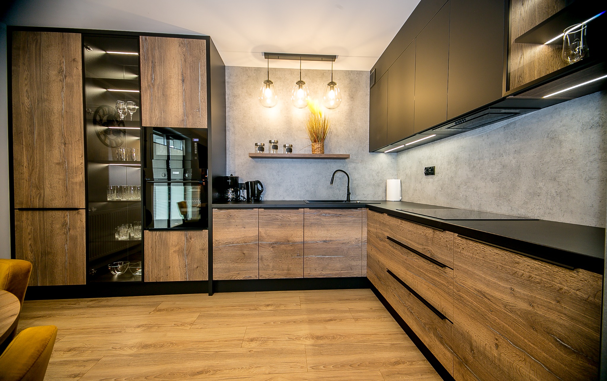 Nowoczesna kuchnia z drewnopodobnymi frontami szafek dolnych i czarnymi szafkami górnymi, oświetleniem LED pod szafkami i trzema wiszącymi lampami z żarówkami edisona, na blacie czajnik i ekspres...