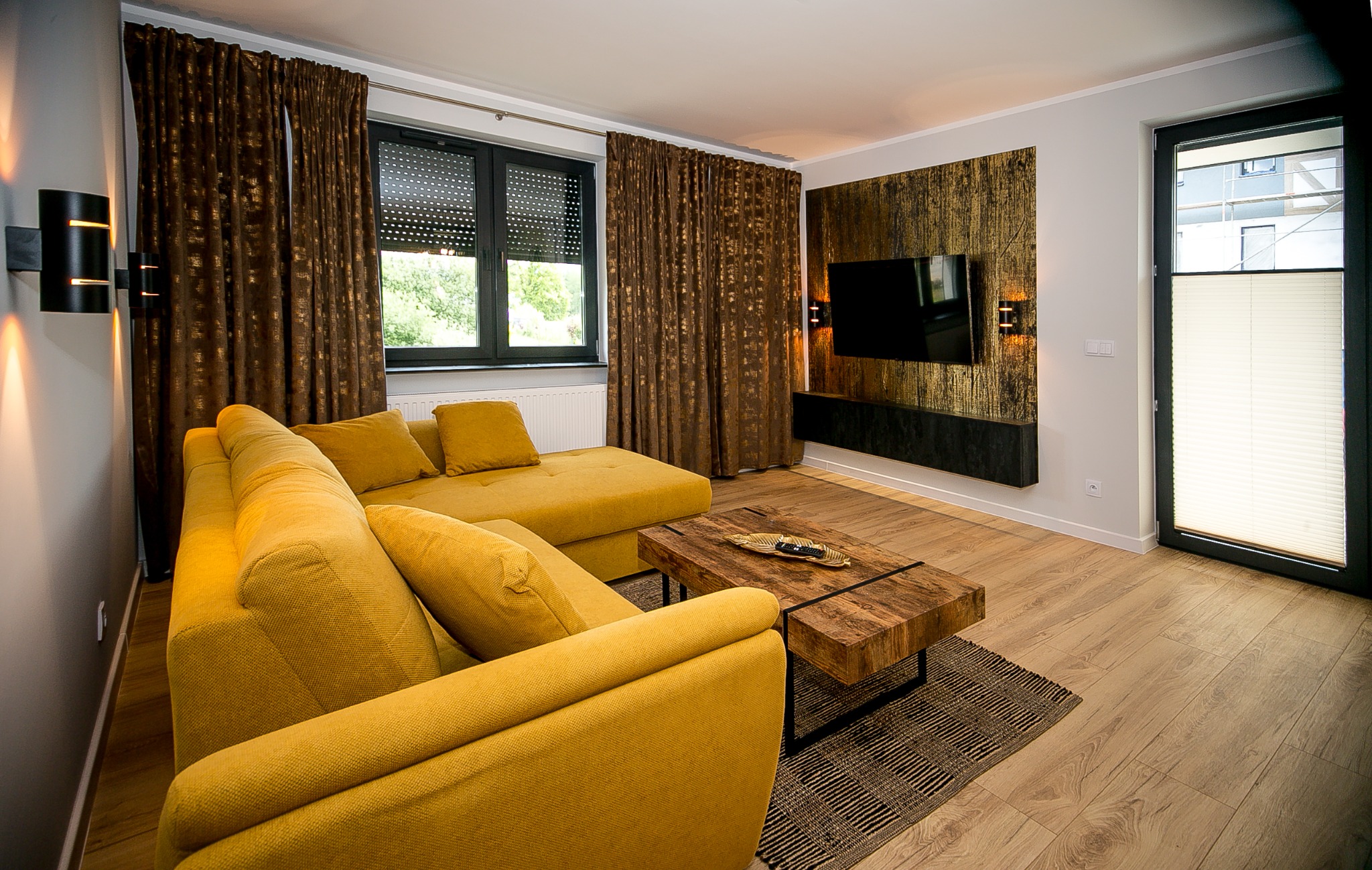 Nowoczesny salon z żółtą sofą narożną, drewnianym stolikiem kawowym na czarnym dywanie i ścianą z dekoracyjną tapetą w odcieniach złota i czerni, na której wisi telewizor.