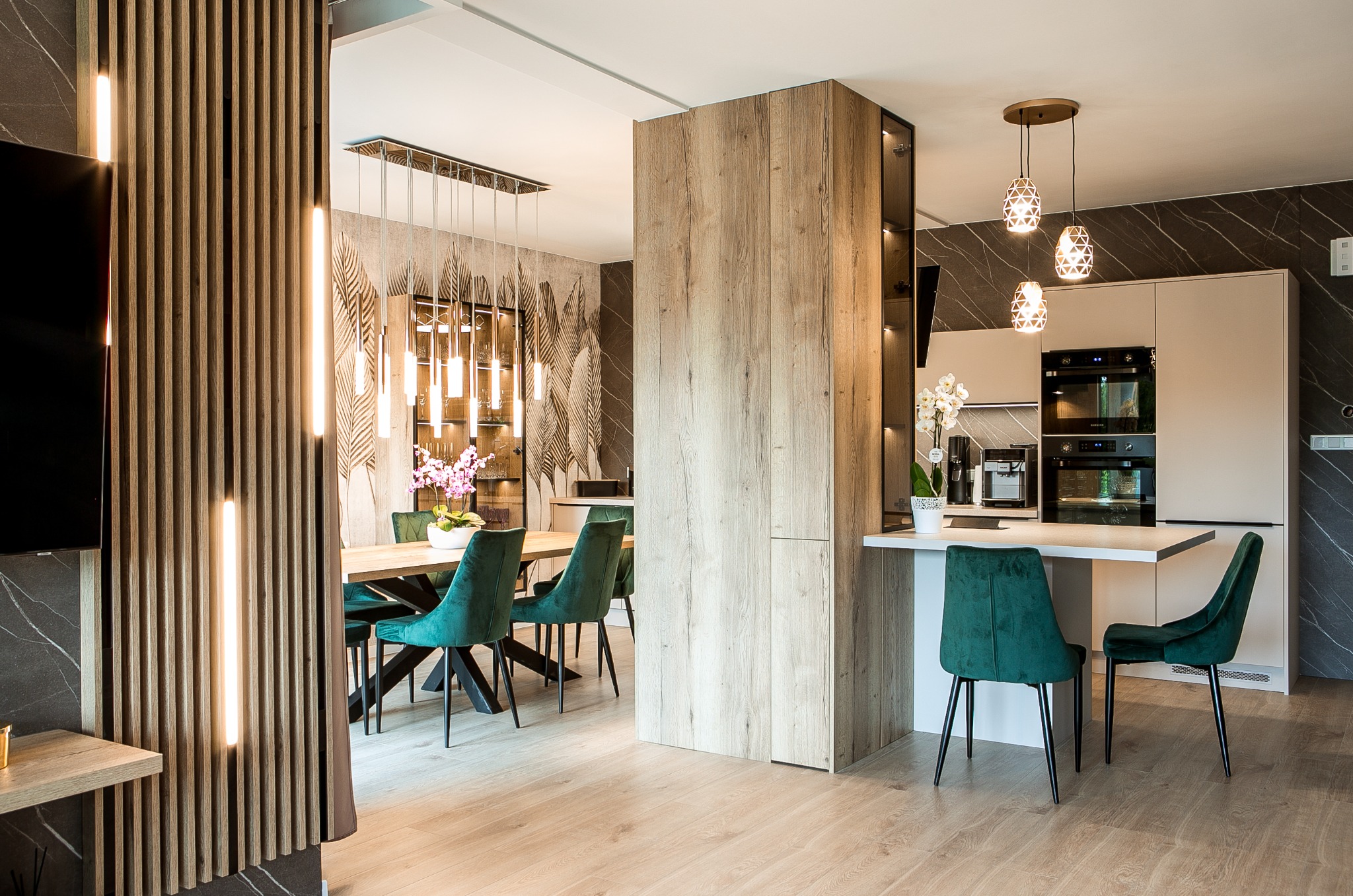 Nowoczesne wnętrze z otwartą kuchnią, jadalnią i salonem, akcentowane przez drewniane panele z oświetleniem LED, marmurowe ściany i smaragdowe krzesła velvetowe przy stole jadalnym, z widoczną...