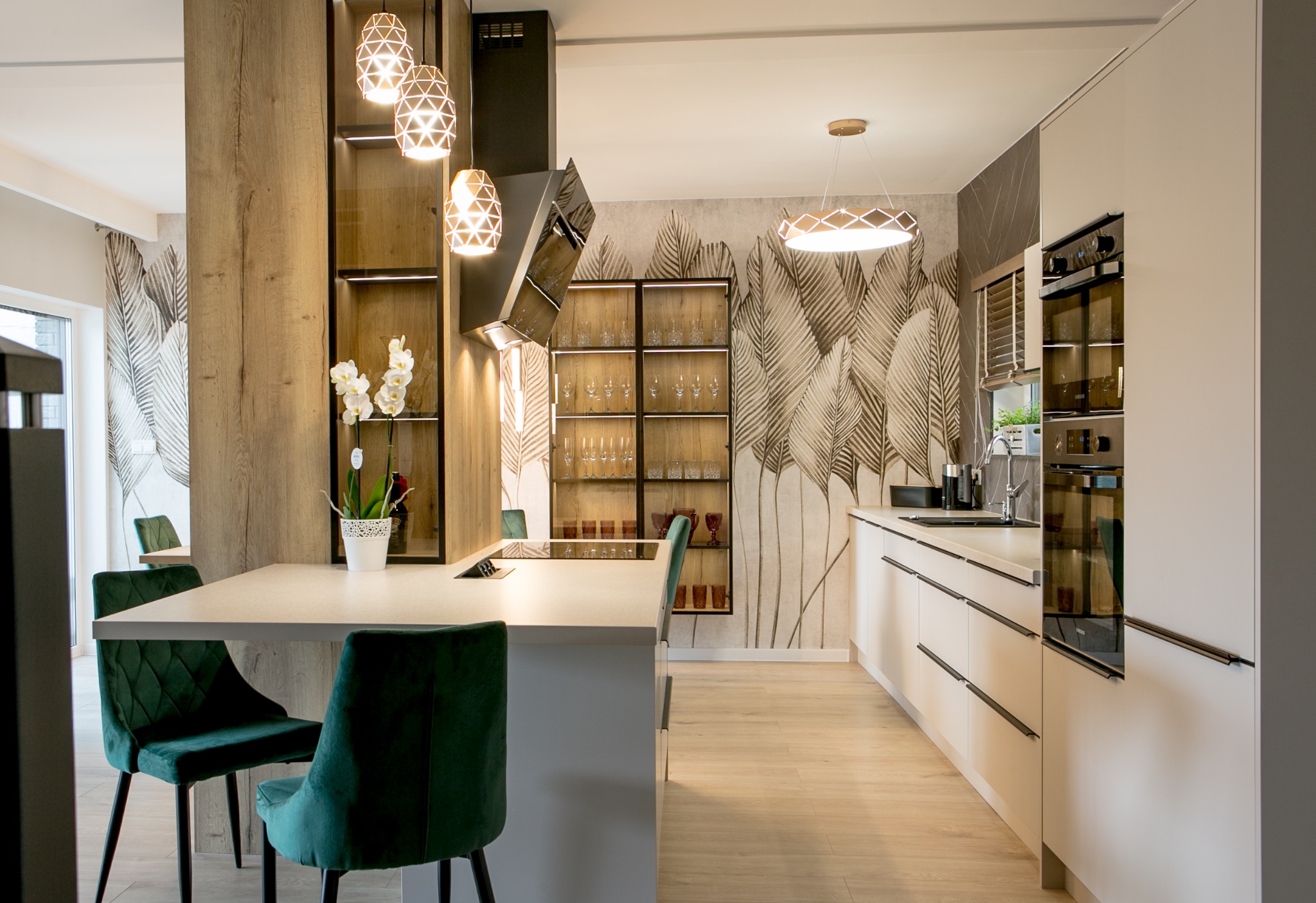 Nowoczesna kuchnia z wyspą, zielonymi krzesłami, białymi szafkami i tapetą w liście, oświetlona wiszącymi lampami o geometrycznych kloszach.