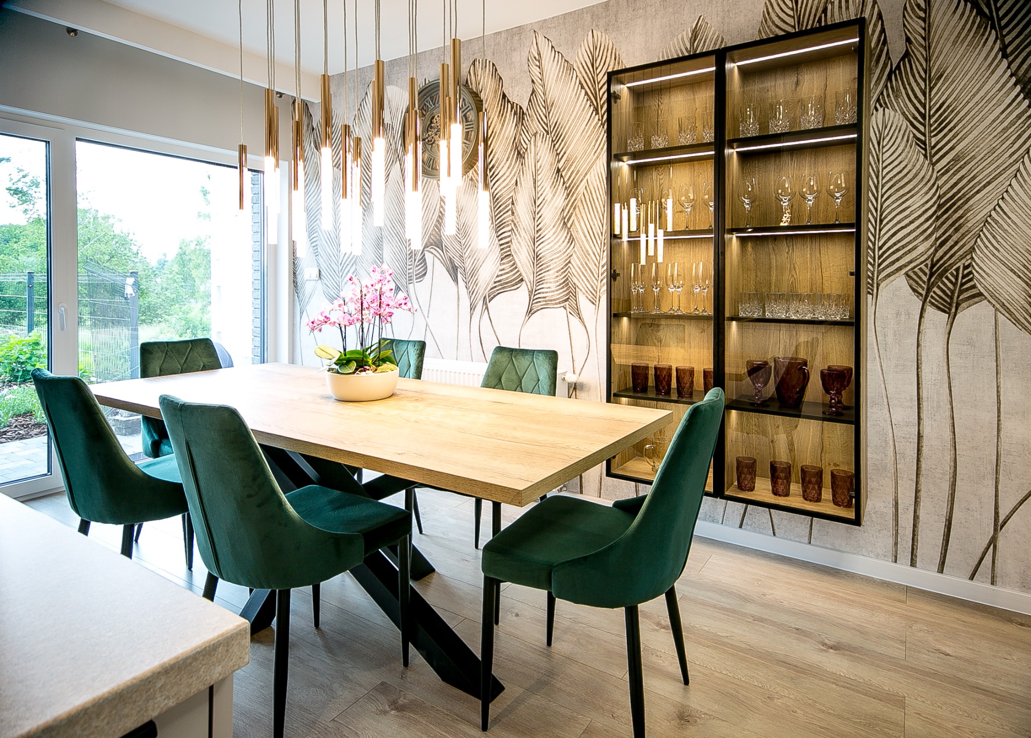 Jadalnia z drewnianym stołem na czarnej podstawie, zielonymi welurowymi krzesłami, wiszącą lampą z mosiężnymi elementami i podświetlaną przeszkloną witryną na tle tapety w liście.