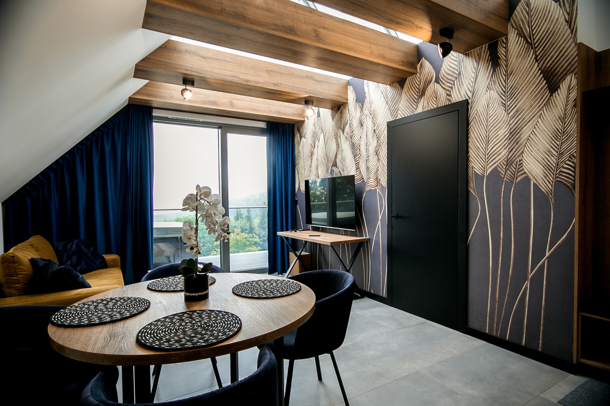Salon w stylu nowoczesnym z drewnianym stołem, czarnymi krzesłami, tapetą w motywy liści i drewnianymi belkami sufitowymi, doświetlony naturalnym światłem z dużego okna balkonowego.