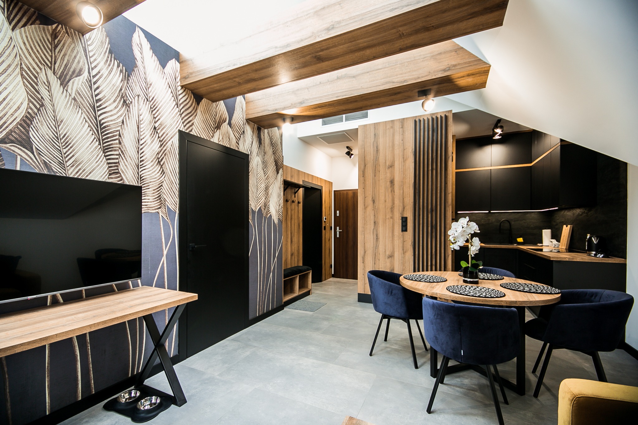 Nowoczesne wnętrze mieszkania z drewnianymi belkami na suficie, tapetą w motywy liści, czarnymi elementami i okrągłym stołem z aksamitnymi krzesłami w kolorze granatowym.