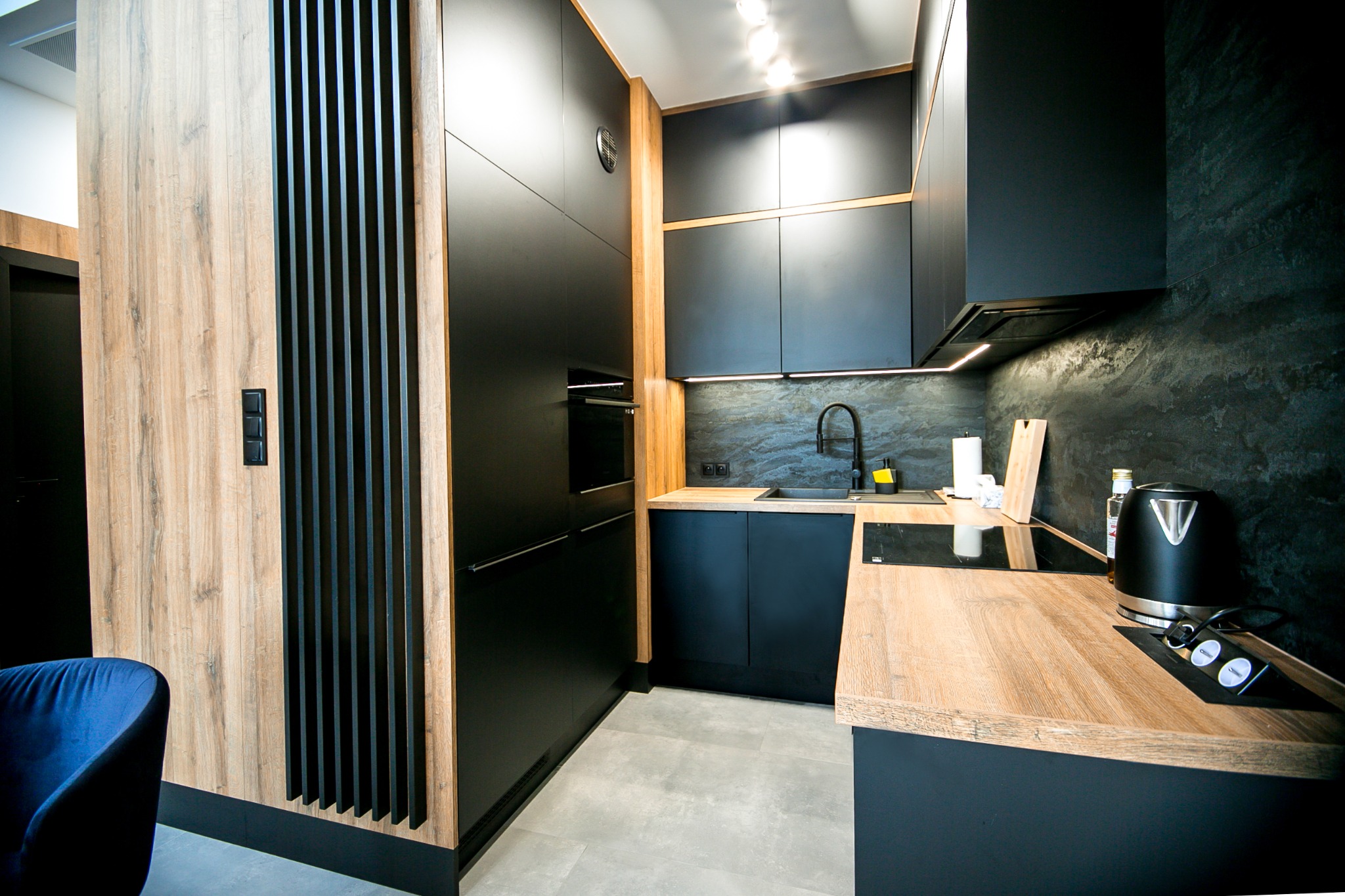 Nowoczesna kuchnia z czarnymi szafkami, drewnianymi blatami i elementami dekoracyjnymi w kolorze drewna, oświetlona punktowymi lampami sufitowymi.