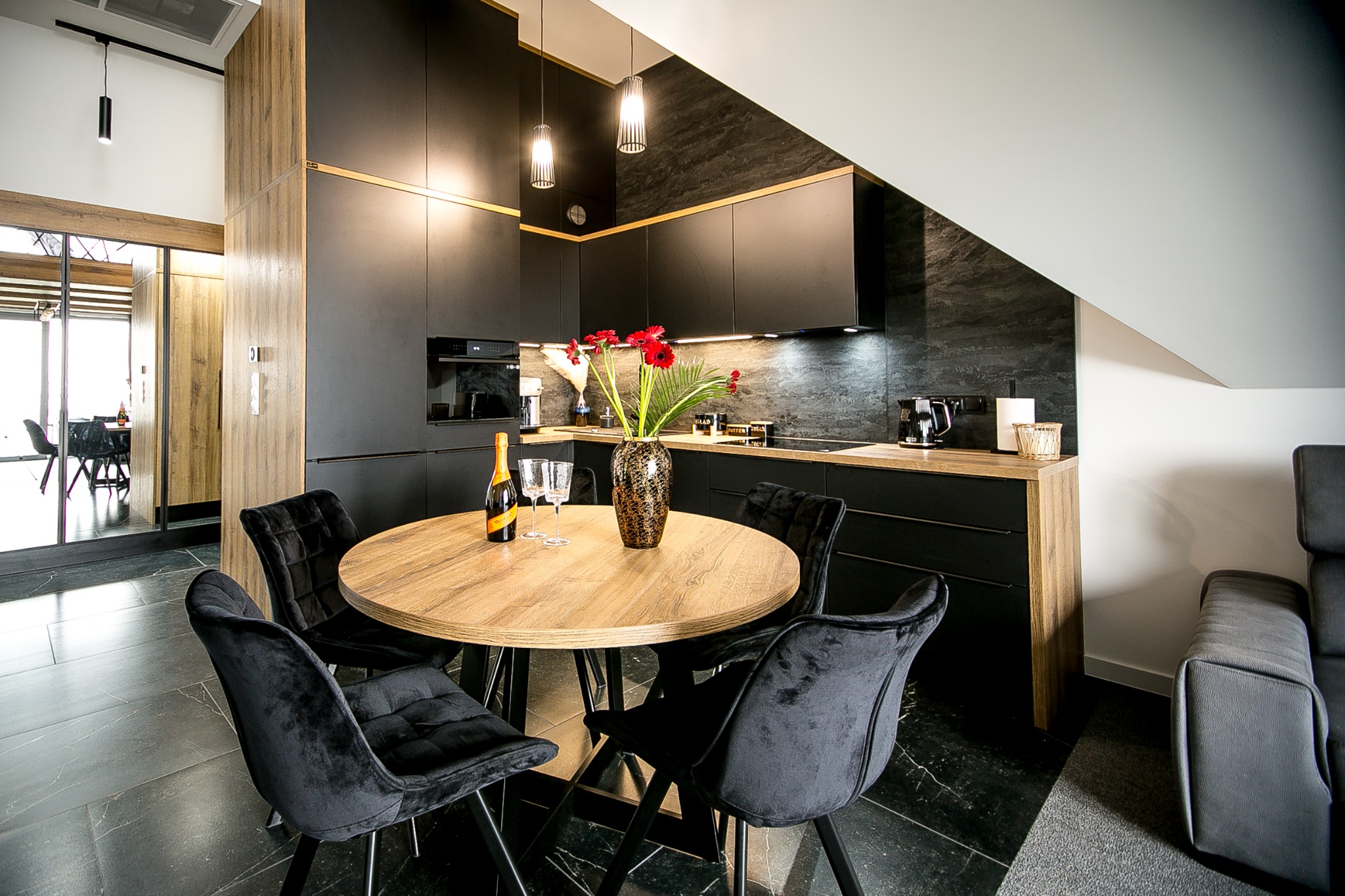 Nowoczesna kuchnia z czarnymi szafkami, drewnianym blatem i okrągłym stołem z czarnymi krzesłami. Na stole wazon z czerwonymi kwiatami, butelka i kieliszki. Widoczne elementy wyposażenia kuchennego.