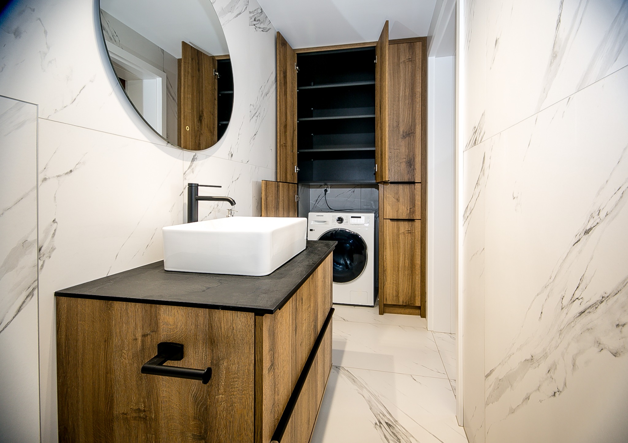 Nowoczesna łazienka z umywalką nablatową, szafką z drewnopodobnym frontem i czarnymi uchwytami, okrągłym lustrem, pralką ukrytą w zabudowie oraz marmurowymi płytkami na ścianach i podłodze.