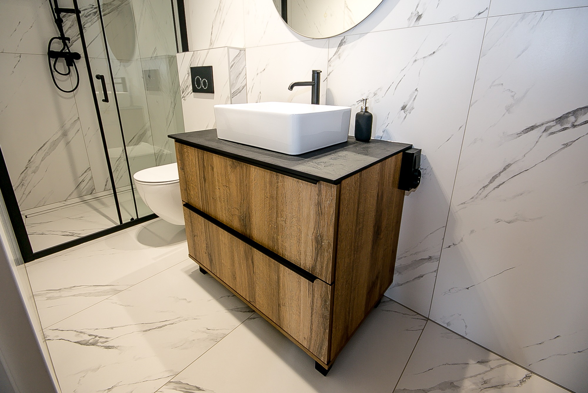Nowoczesna łazienka z białą umywalką nablatową na drewnopodobnej szafce z czarnym blatem, czarna armatura, marmurowe płytki na ścianach i podłodze, kabina prysznicowa w tle.