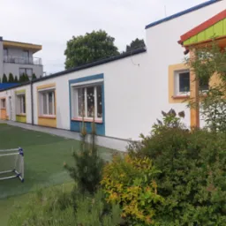 Kolorowa elewacja budynku z oknami o różnych barwach obramowań, otoczona zielenią, w tle budynek mieszkalny.