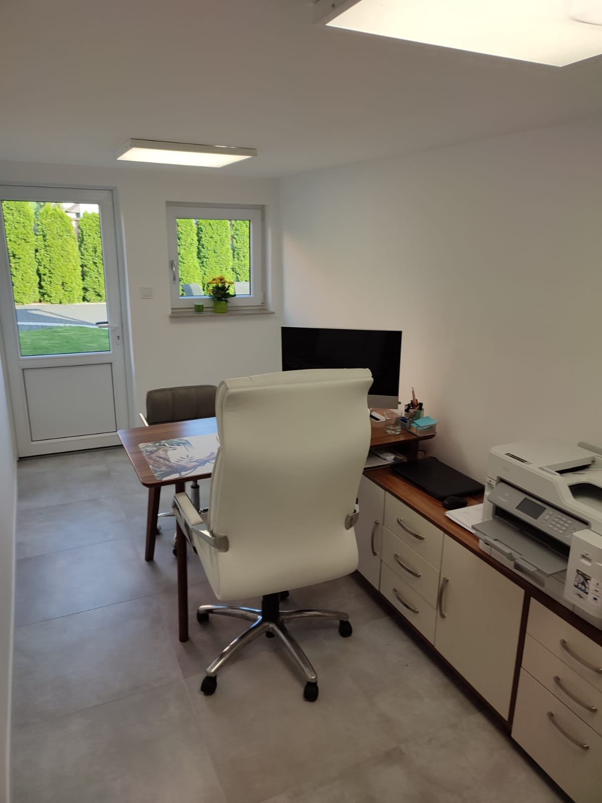 Jasne biuro z biurkiem, krzesłem obrotowym, monitorem, drukarką i widokiem na zadbany ogród przez okno i drzwi balkonowe.