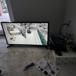 Radosław Sekuła Elektro RS - Monitor w Składzistem wyświetla obraz z kamery monitoringu zewnętrznego w zimowej scenerii, widoczne kable, router i instrukcja obsługi systemu alarmowego.