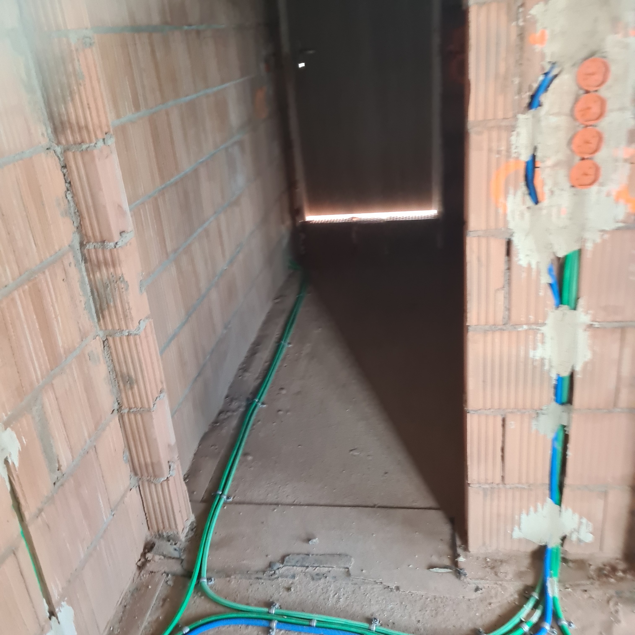 Wnętrze budynku w trakcie budowy, widoczne kable elektryczne w kolorze zielonym i niebieskim, przymocowane do ściany z cegły dziurawki, oraz puszki instalacyjne zalane gipsem.