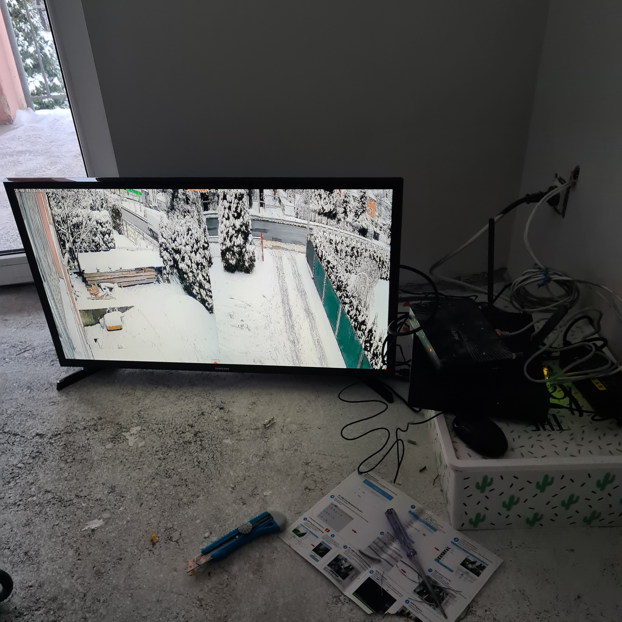 Monitor w Składzistem wyświetla obraz z kamery monitoringu zewnętrznego w zimowej scenerii, widoczne kable, router i instrukcja obsługi systemu alarmowego.