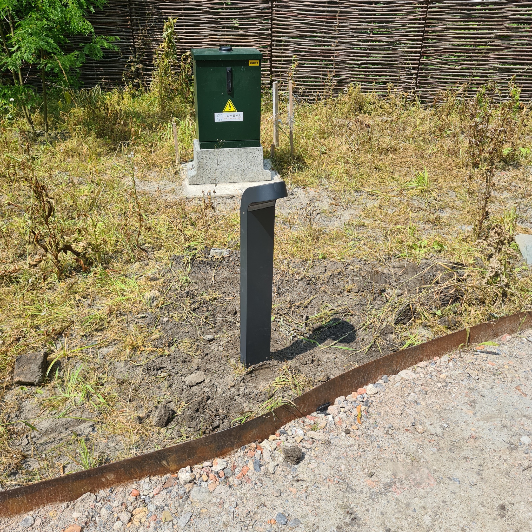 Zewnętrzna szafka elektryczna z żółtym symbolem ostrzegawczym na betonowej podstawie, obok nowoczesnej, minimalistycznej lampy ogrodowej, na tle wiklinowego płotu i zarośniętego terenu.