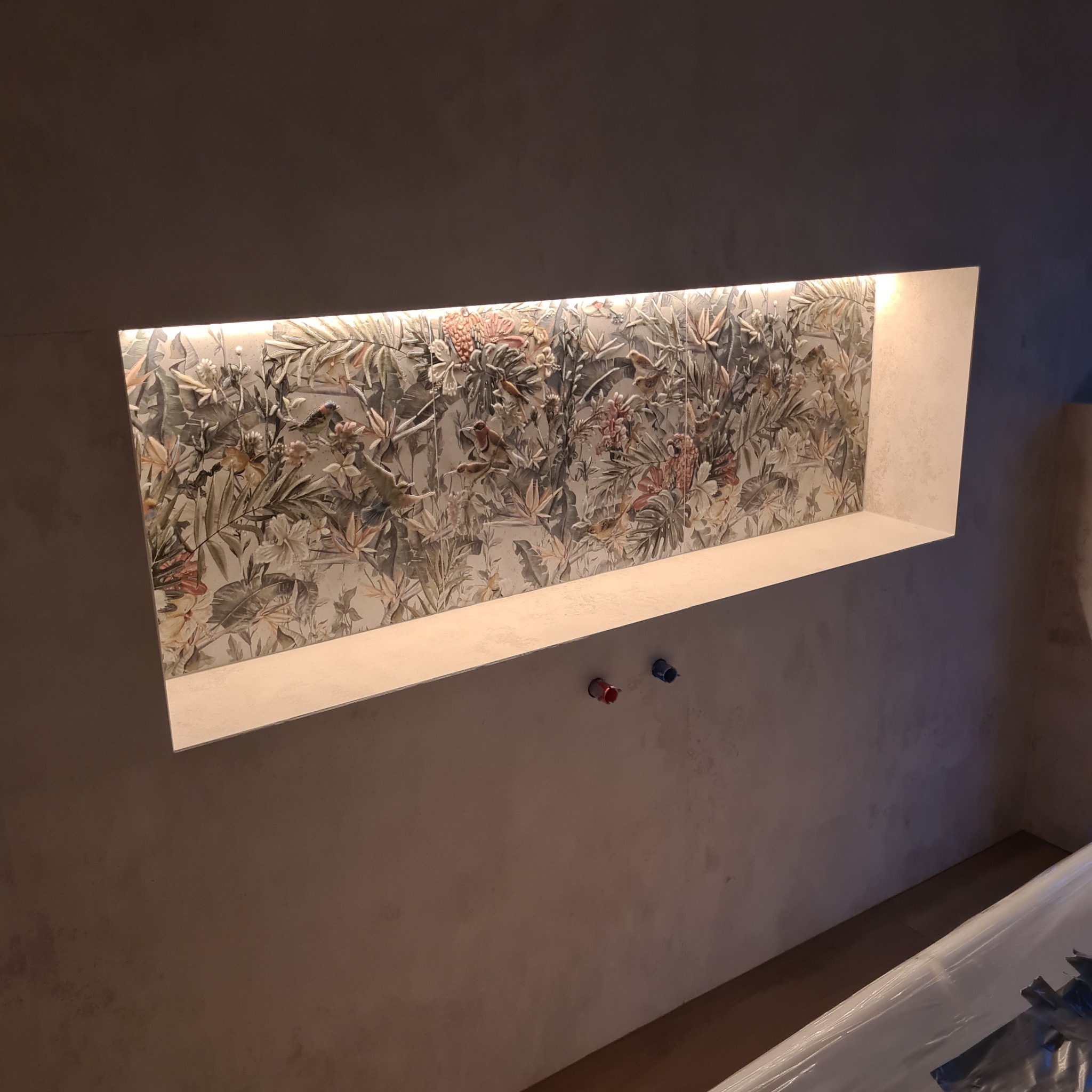 Wnęka w ścianie z podświetlaną, dekoracyjną płytką ceramiczną o motywie roślinnym i ptasim, widoczne rury instalacji wodnej.