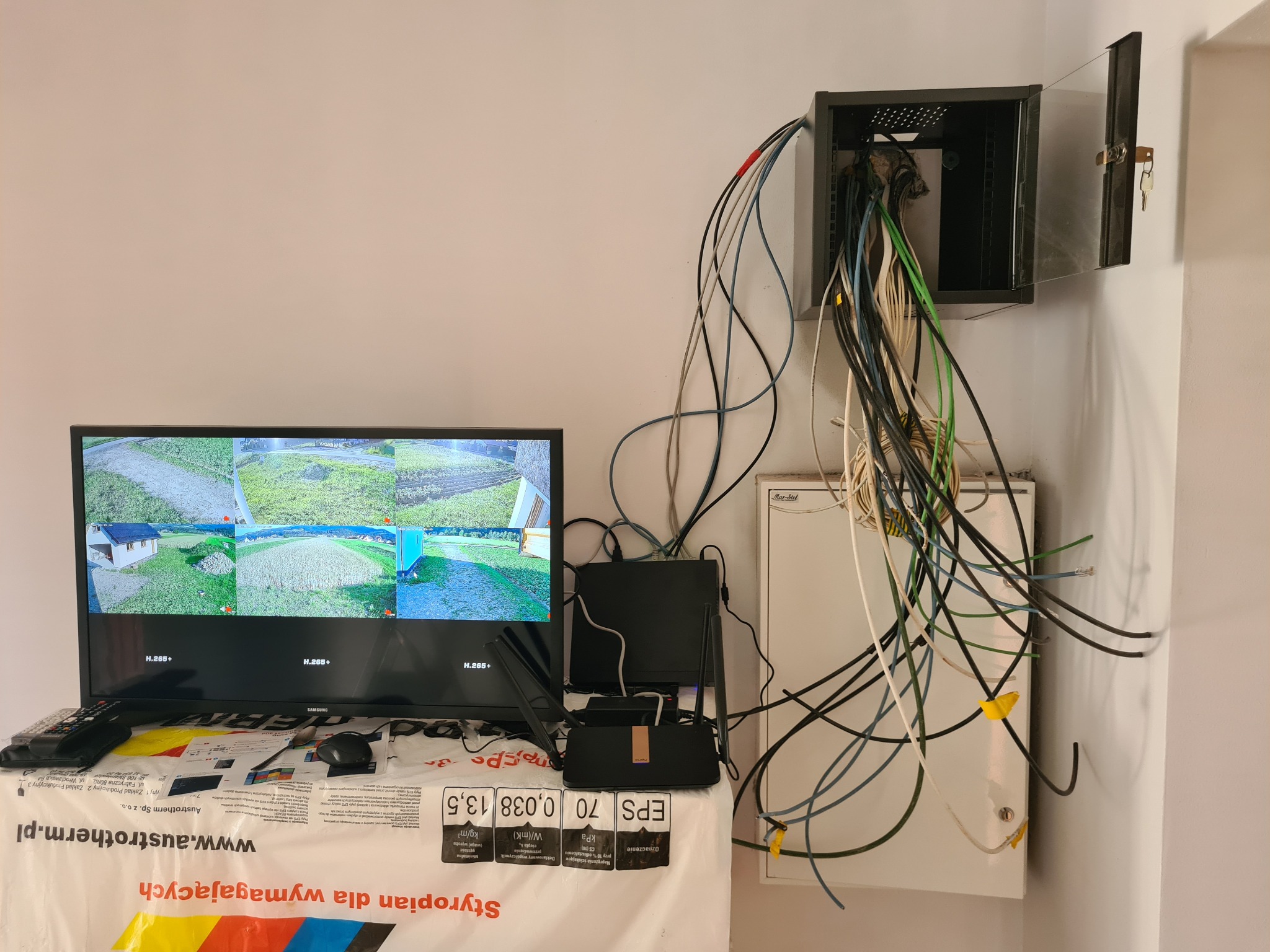 System monitoringu z widokiem z kamer na ekranie telewizora, router i skrzynka z kablami na tle ściany.