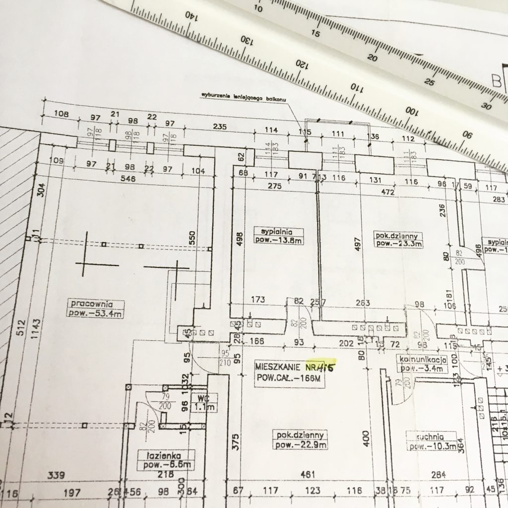 Plan mieszkania z naniesionymi wymiarami i układem pomieszczeń, częściowo zakryty przez białą linijkę. Widoczne opisy pomieszczeń i ich powierzchni.