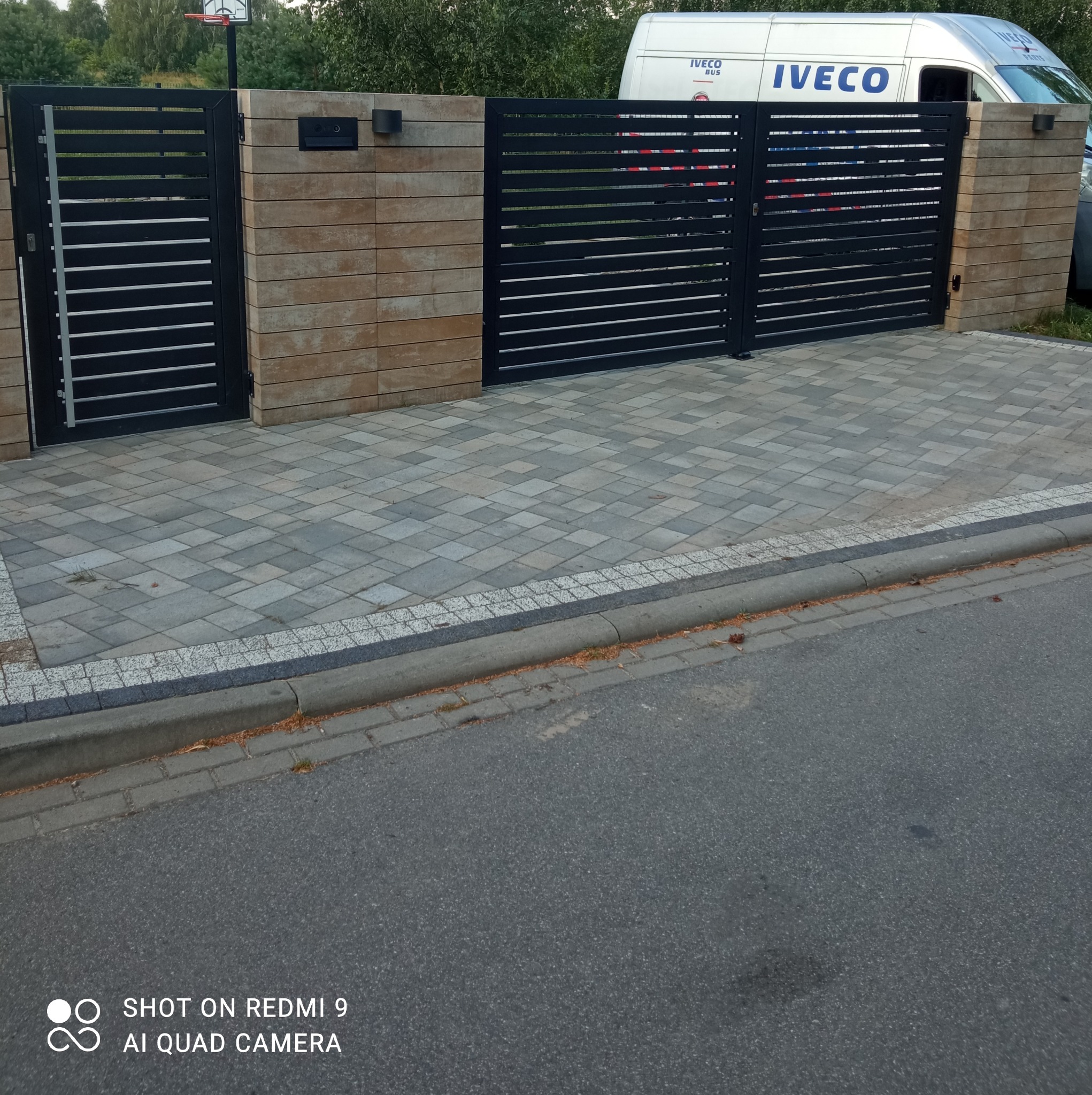 Czarna, pozioma brama panelowa z jasnymi przerwami, osadzona w murze z beżowych bloczków, na posesji wyłożonej szarą kostką brukową, z białym samochodem dostawczym IVECO w tle.