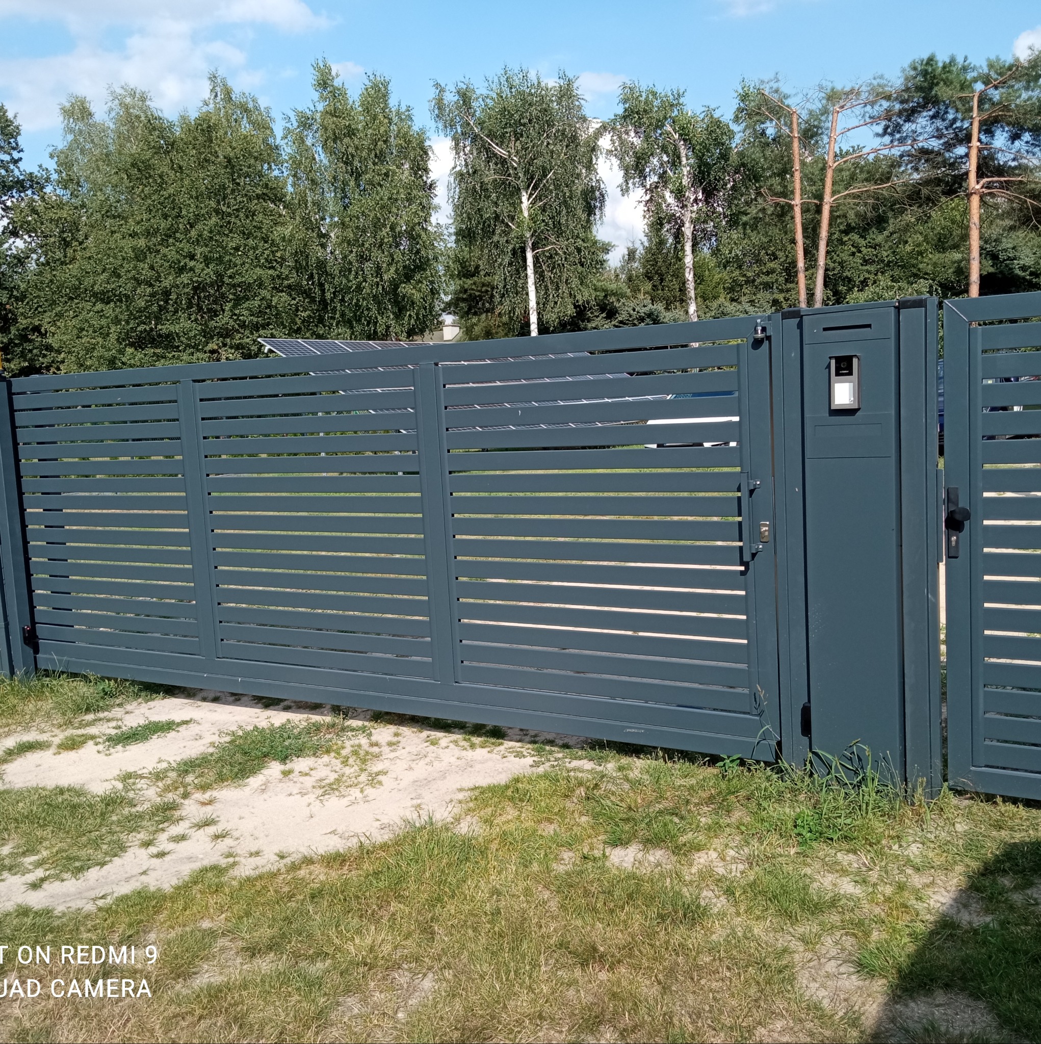 Nowoczesna, szara brama przesuwna z poziomymi panelami i wideodomofonem, na tle zieleni i drzew