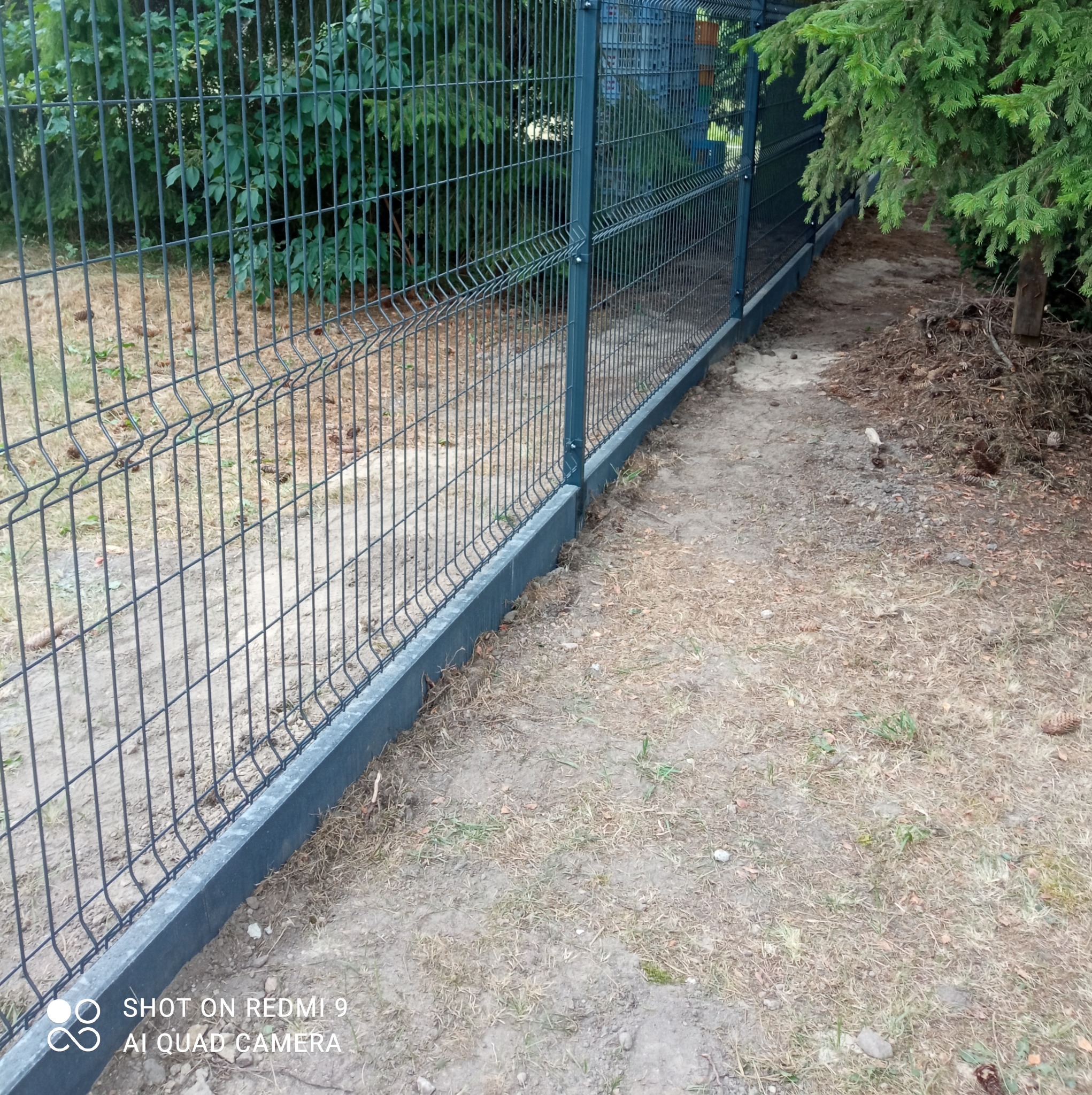 Szary panelowy płot z podmurówką w Łasku, widok z boku, z fragmentem trawnika i drzew w tle.