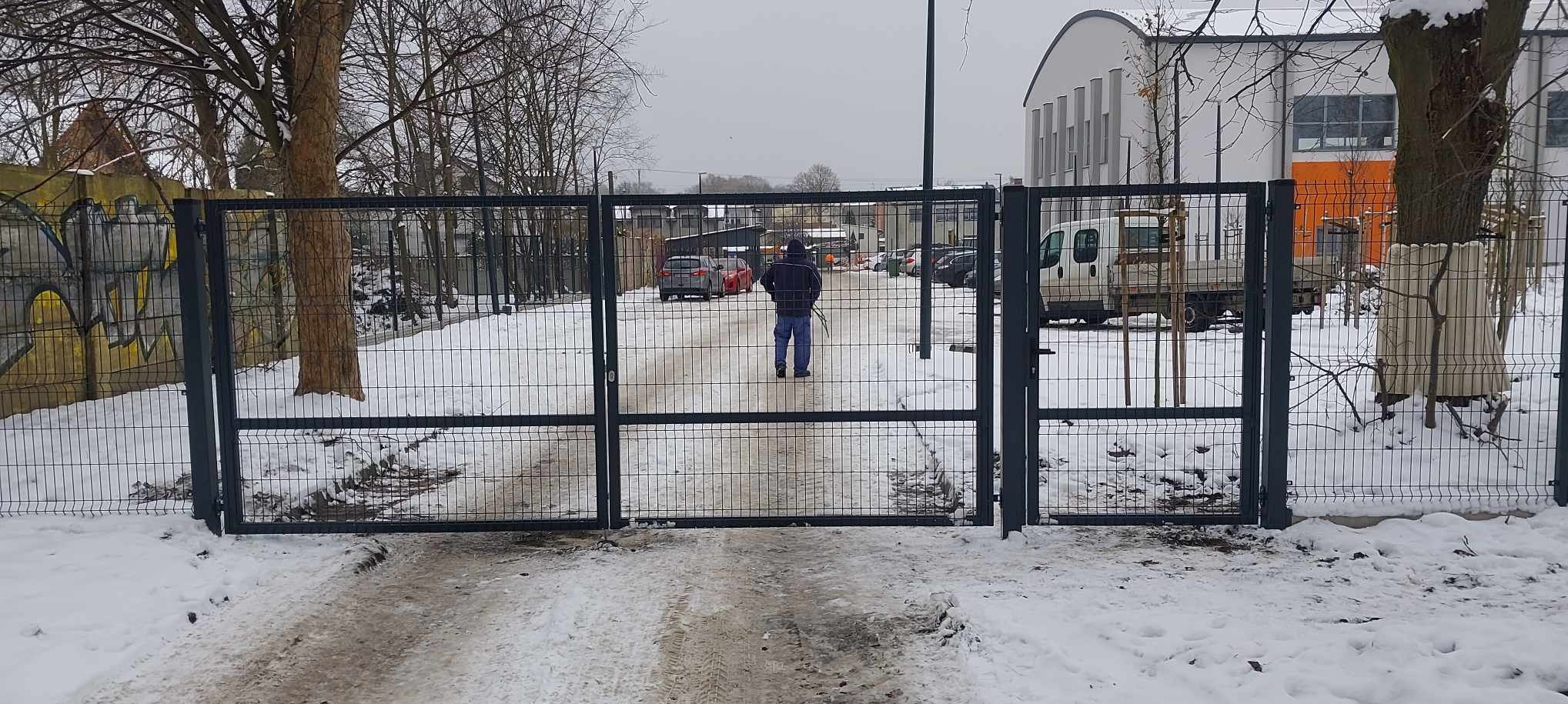 Otwarta, ciemna brama panelowa z widokiem na zasnieżony podjazd i budynek w tle, mężczyzna oddala się w głąb posesji.