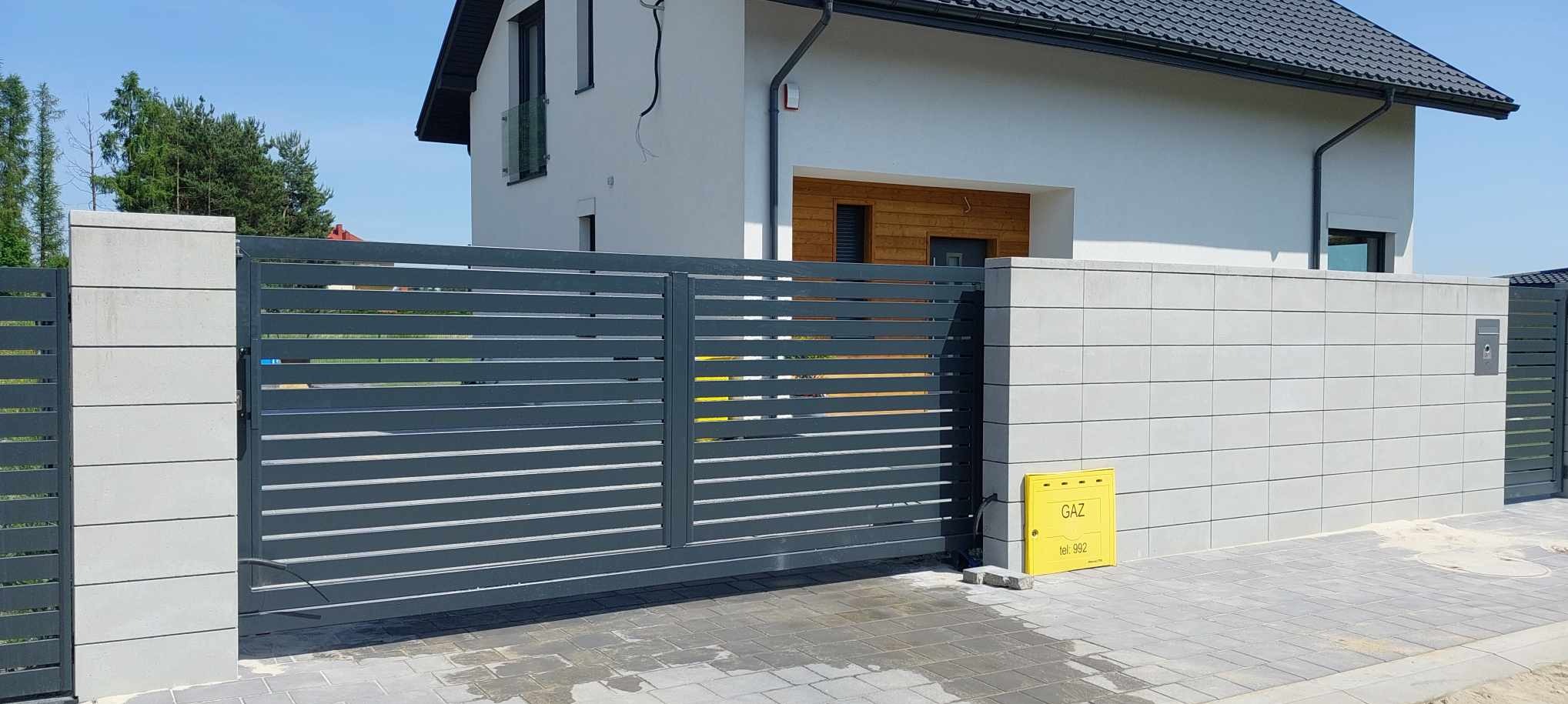 Nowoczesna, przesuwna brama wjazdowa z poziomych profili, w kolorze szarym, zamontowana przy ogrodzeniu z bloczków betonowych. Widoczna skrzynka gazowa w kolorze żółtym.