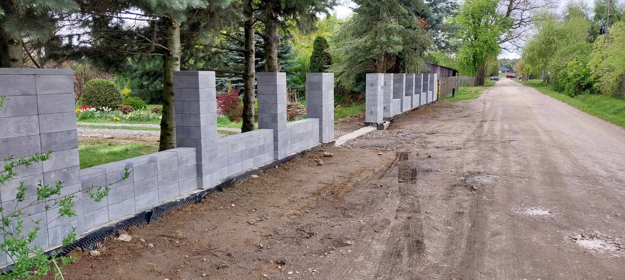 Wznoszenie ogrodzenia z szarych, betonowych bloczków modułowych z widoczną membraną izolacyjną przy gruncie, na tle wiejskich zabudowań i drzew.