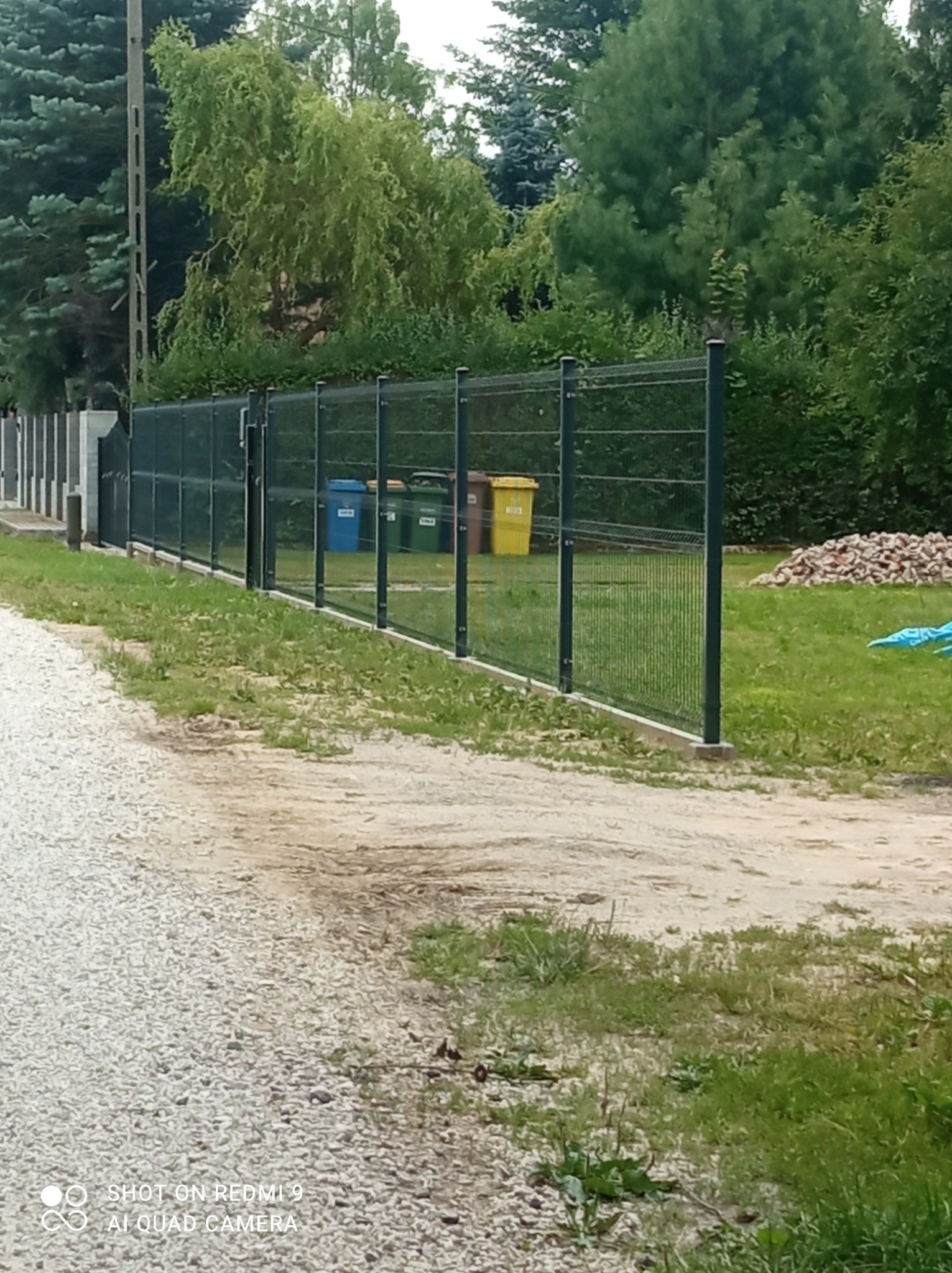Zielone ogrodzenie panelowe z widocznymi trzema pojemnikami na śmieci w tle, z perspektywy ścieżki żwirowej.