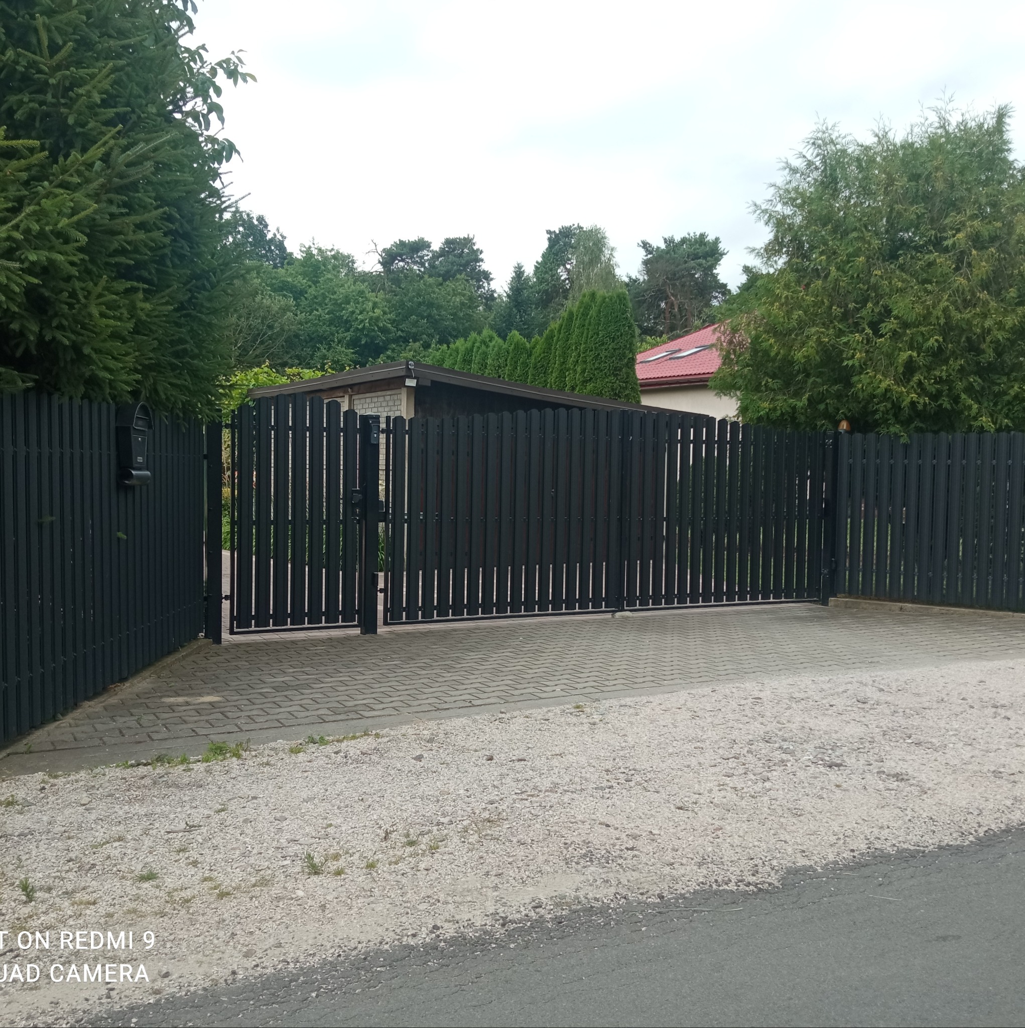 Czarna, metalowa brama wjazdowa i furtka z pionowych profili, prowadząca na posesję z kostką brukową i żwirowym podjazdem, otoczona zielenią drzew i krzewów.