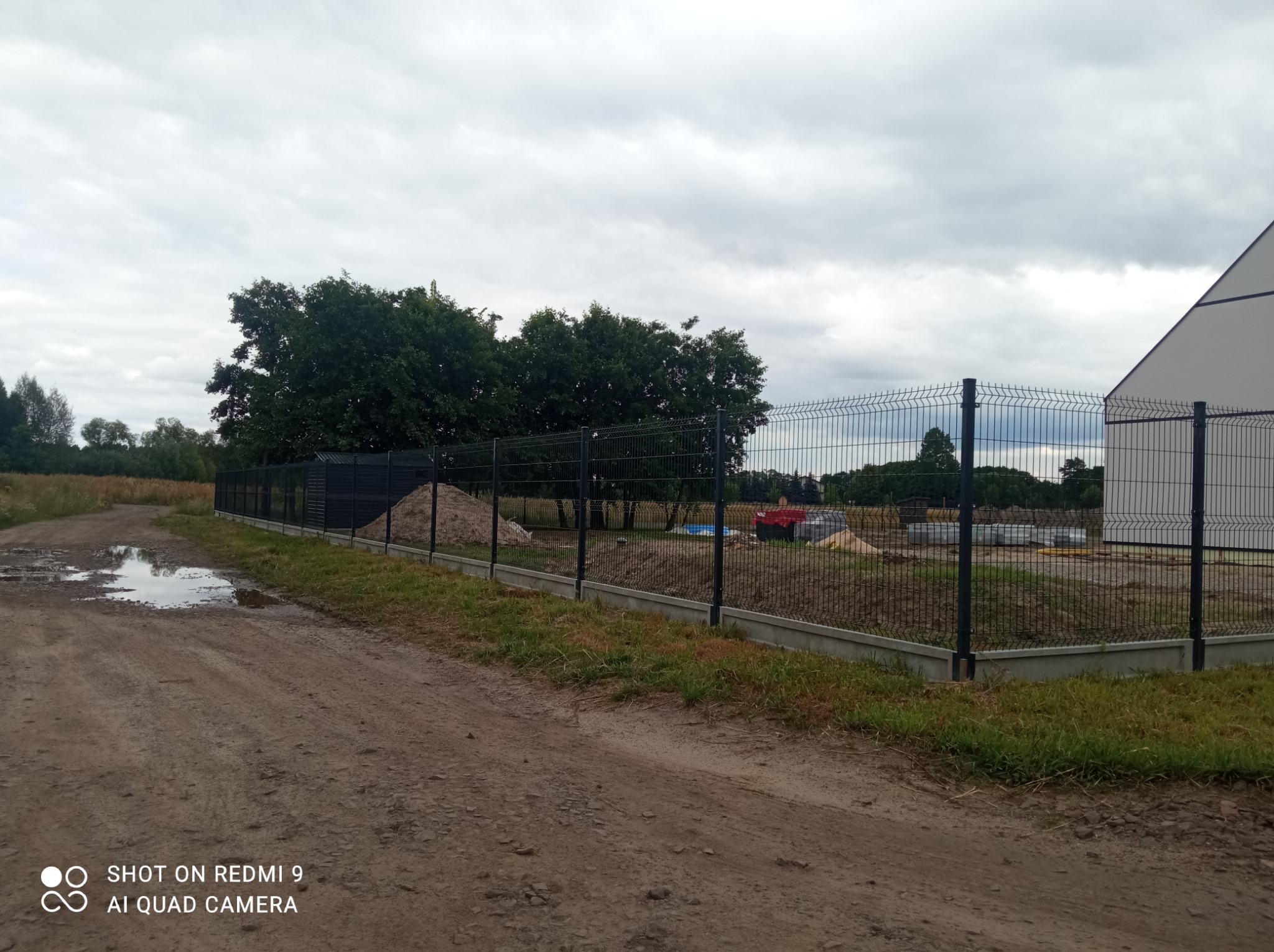 Ciemnoszare, metalowe ogrodzenie panelowe z podmurówką betonową, widoczne z polnej drogi z kałużami, na tle budowy i pochmurnego nieba.