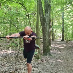 Błażej Wit BWIT - Mężczyzna z brodą, w lesie, rozciąga mięśnie ramion za pomocą kijków do nordic walking, przygotowując się do biegu.