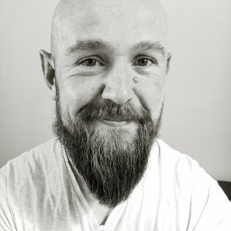 Błażej Wit BWIT - Portret łysiejącego mężczyzny z brodą, uśmiechającego się, z pieprzykiem na skroni, w białym T-shircie, na jasnym tle.