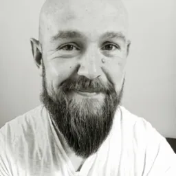 Portret łysiejącego mężczyzny z brodą, uśmiechającego się, z pieprzykiem na skroni, w białym T-shircie, na jasnym tle.