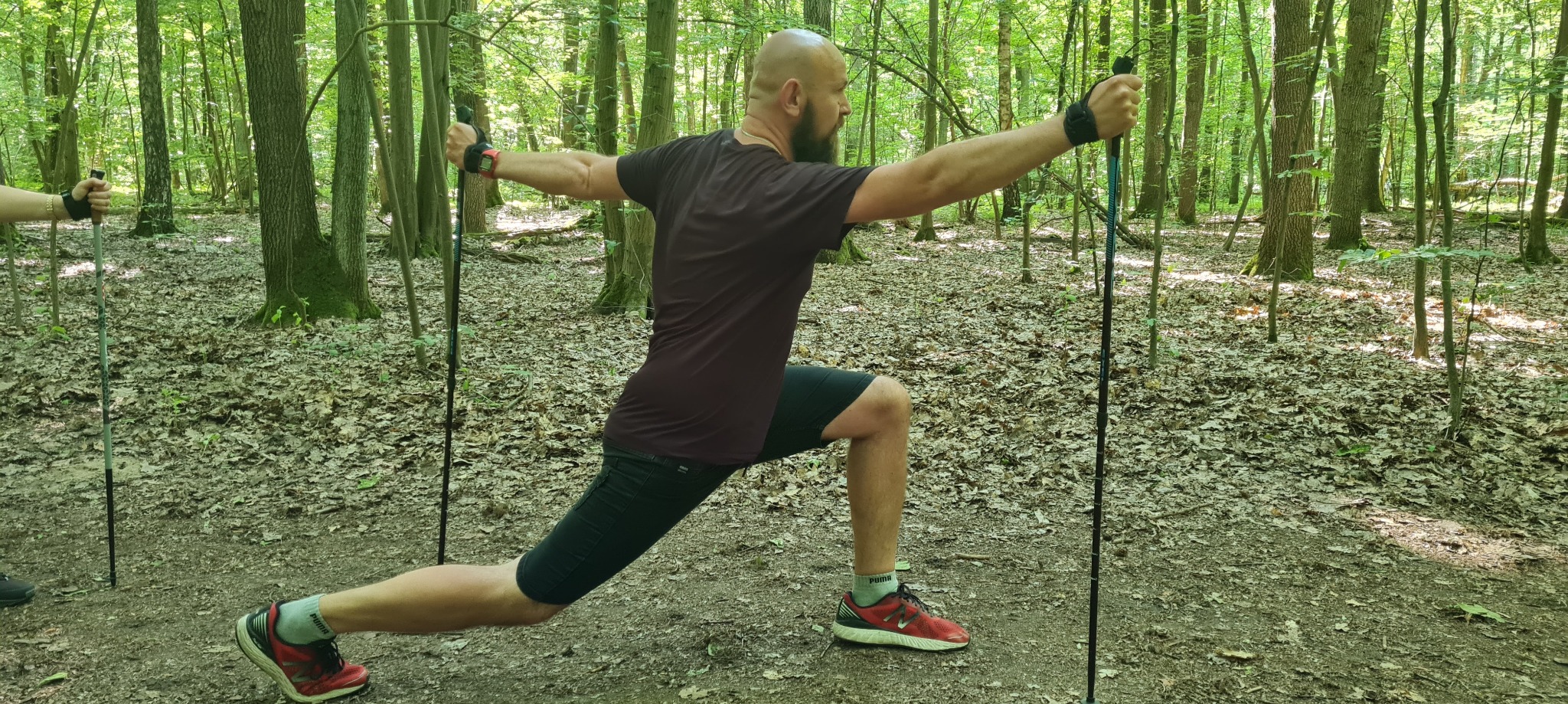 Mężczyzna z brodą wykonuje wypad w przód z kijkami do nordic walking w lesie, rozciągając ramiona na boki, widoczne czerwone buty sportowe Puma i drugi uczestnik z kijkami w tle.