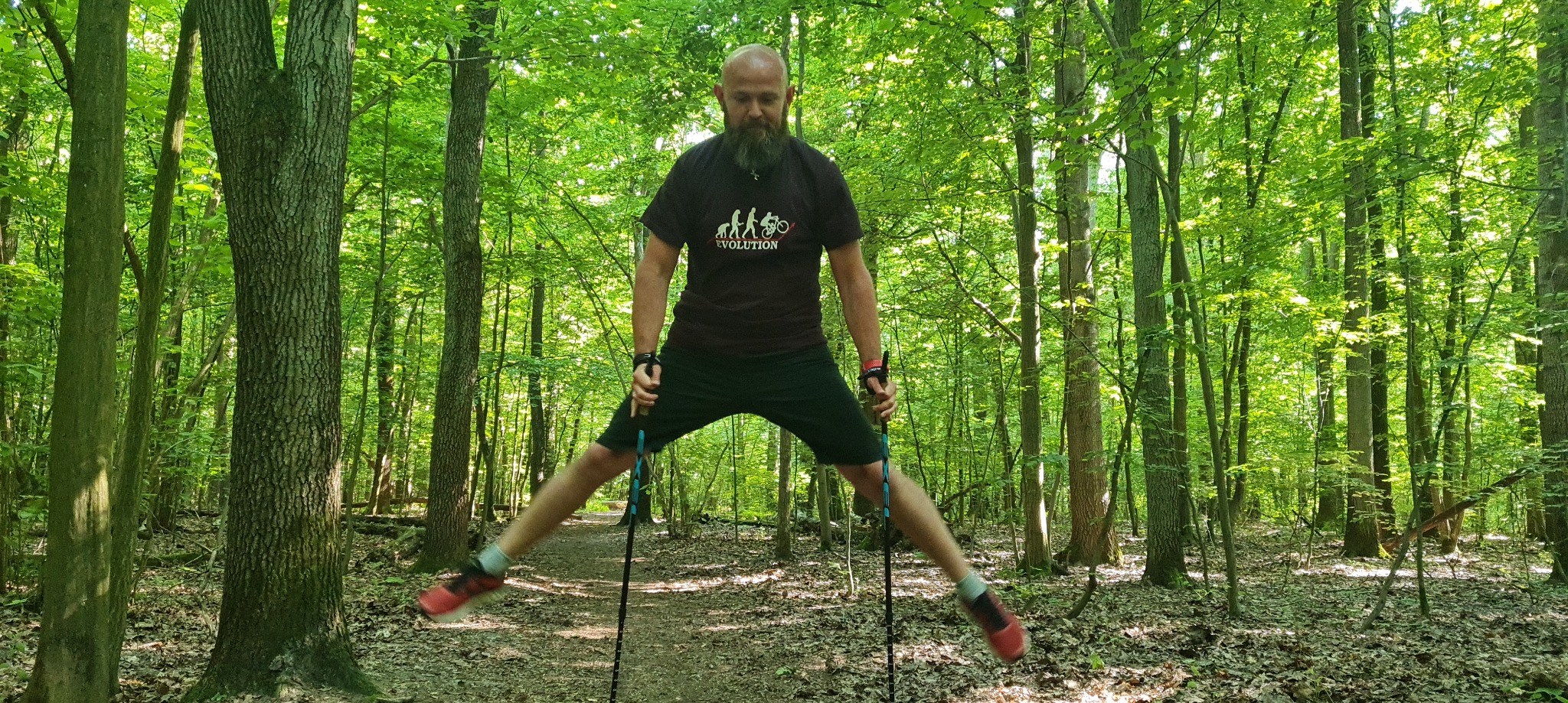 Mężczyzna z brodą w lesie, w rozkroku, używający kijków trekkingowych podczas ćwiczeń rozciągających.