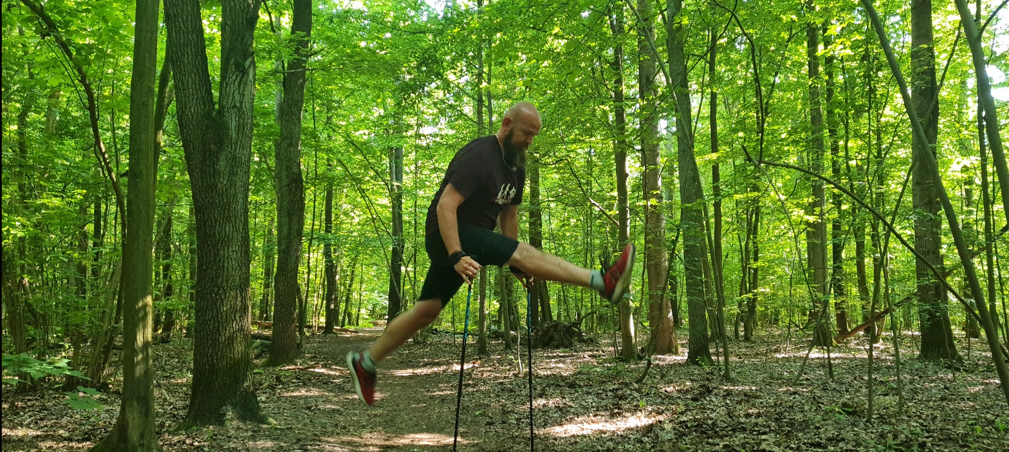 Mężczyzna z brodą w czarnej koszulce i spodenkach, w czerwonych butach, skacze w lesie trzymając kijki do nordic walking.