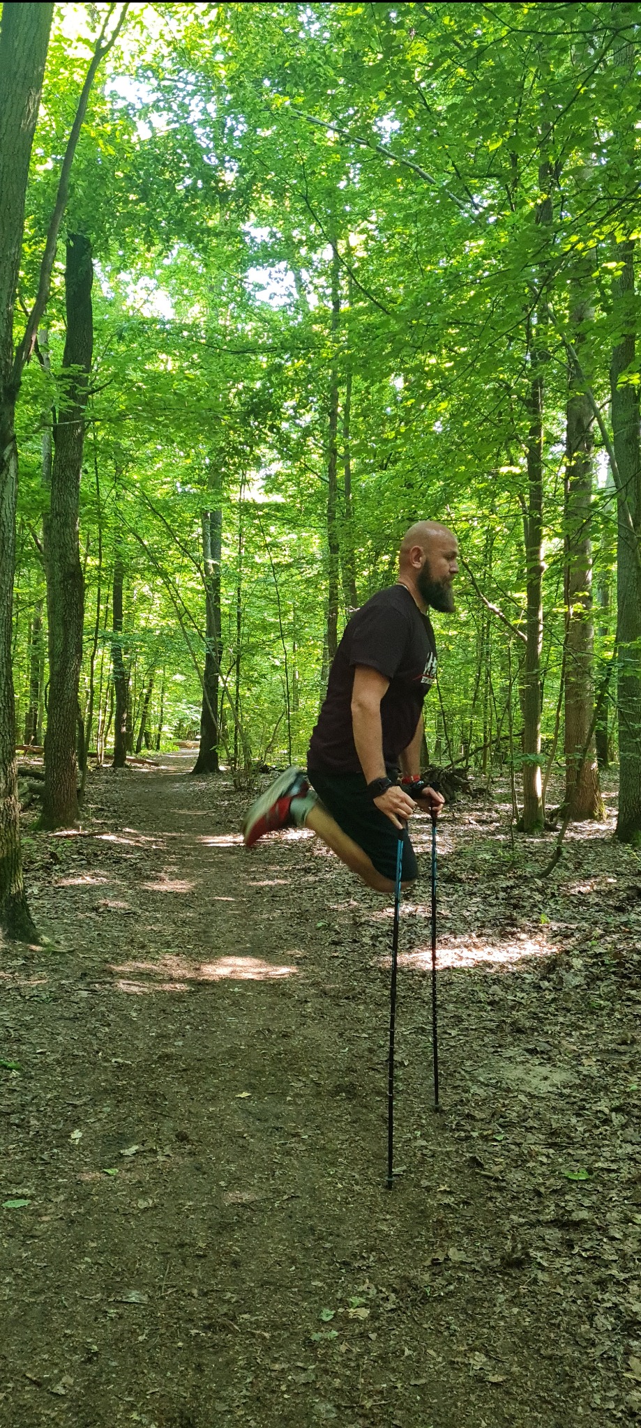 Mężczyzna z brodą w czarnej koszulce i sportowych butach skacze na leśnej ścieżce, trzymając w dłoniach kijki do nordic walking.