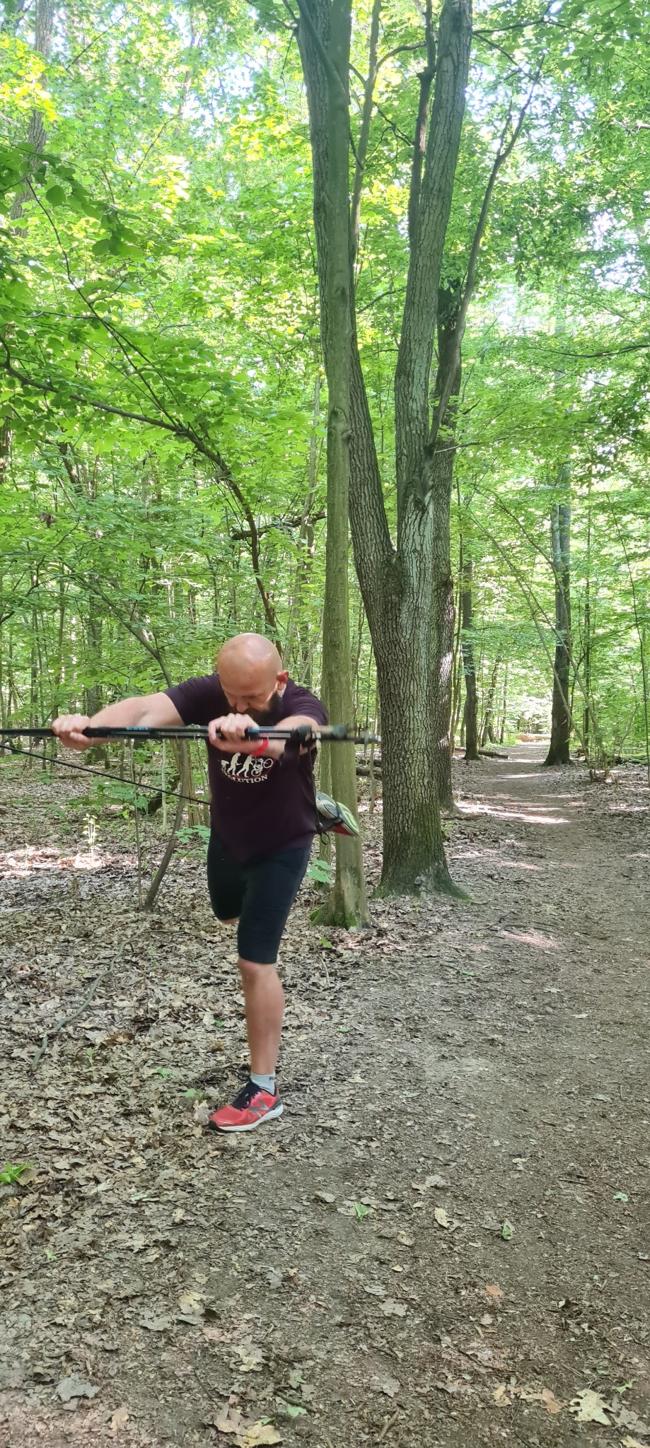 Mężczyzna z brodą, w lesie, rozciąga mięśnie ramion za pomocą kijków do nordic walking, przygotowując się do biegu.