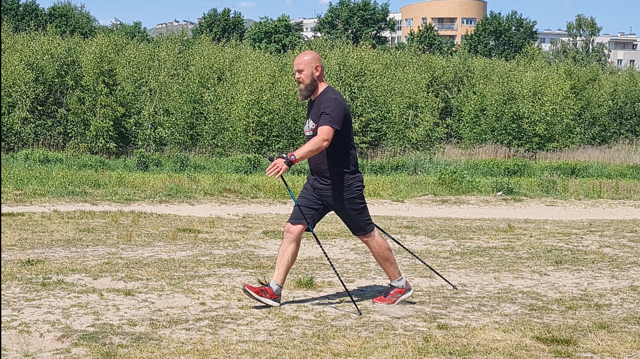 Mężczyzna z brodą, w czarnym stroju i czerwonych butach, uprawia nordic walking na piaszczystym terenie z zielenią w tle, używając kijków z niebieskimi akcentami.