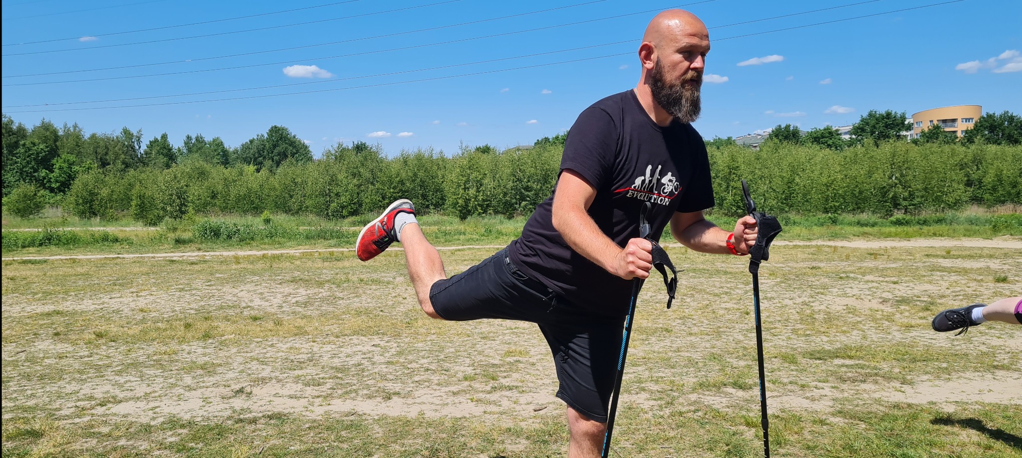 Mężczyzna z brodą w czarnej koszulce z nadrukiem, stojący na trawiastym terenie z kijkami do nordic walking, unosi nogę do tyłu, wykonując rozciąganie w słoneczny dzień.