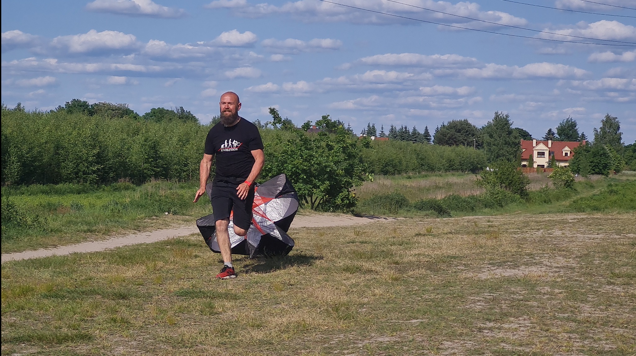 Mężczyzna z brodą w czarnej koszulce z nadrukiem biegnie po trawiastym terenie ciągnąc za sobą zwinięty latawiec treningowy, w tle drzewa i dom.