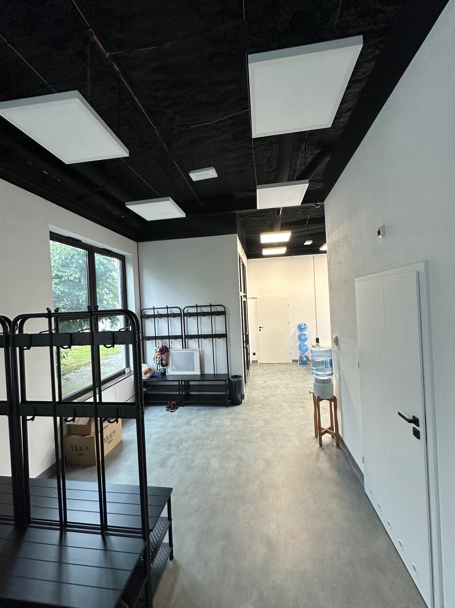 Wnętrze biura z czarnym sufitem, na którym zamontowano podwieszane, kwadratowe panele oświetleniowe LED. Widoczny wieszak na ubrania, okno z widokiem na zieleń oraz dystrybutor wody.