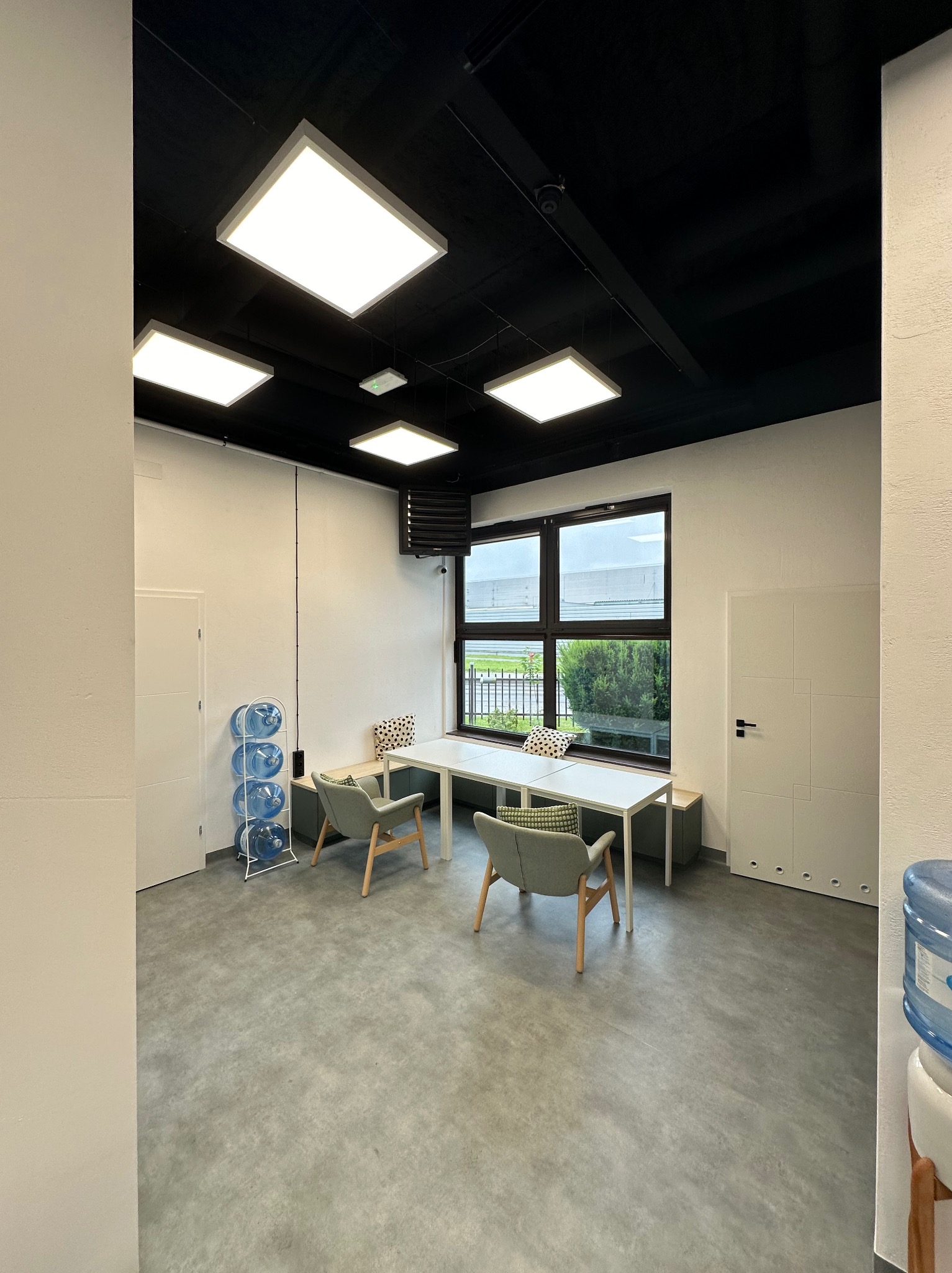 Jasne, nowoczesne biuro z białymi ścianami, czarnym sufitem i geometrycznymi lampami LED. Widoczne dwa krzesła, biały stół, okno z widokiem na zewnątrz oraz dystrybutory wody.