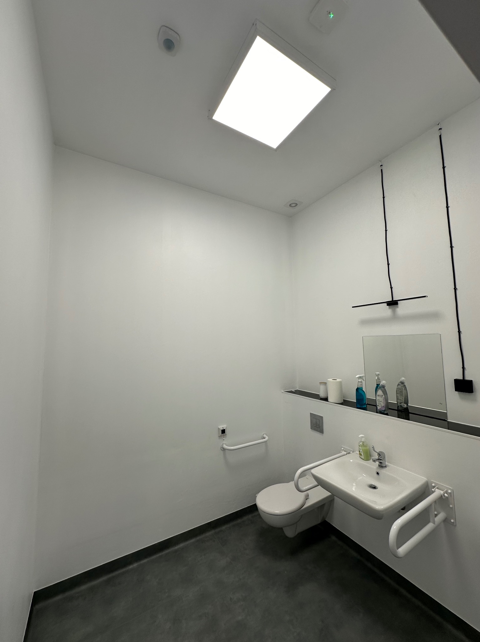 Nowoczesna łazienka w minimalistycznym stylu z umywalką i toaletą dla osób niepełnosprawnych, oświetlona górnym panelem LED i lampą nad lustrem z widocznymi przewodami elektrycznymi.