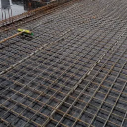 Zbrojenie płyty fundamentowej z widocznymi deskowaniami i przygotowaniami do zalania betonem, w tle zabudowa jednorodzinna w zimowej scenerii.