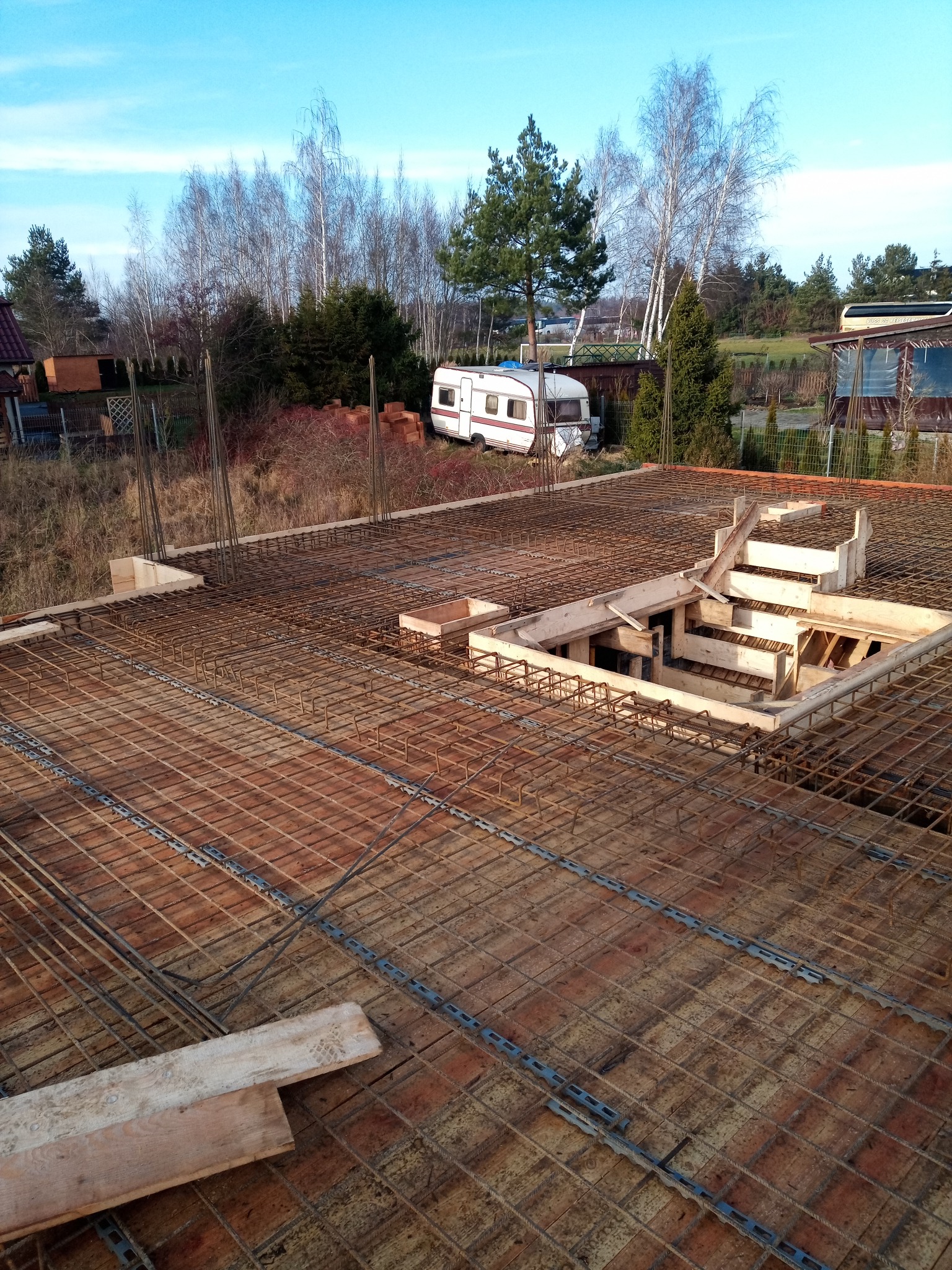 Zbrojenie płyty fundamentowej z drewnianym szalunkiem otworu, w tle drzewa i przyczepa kempingowa.