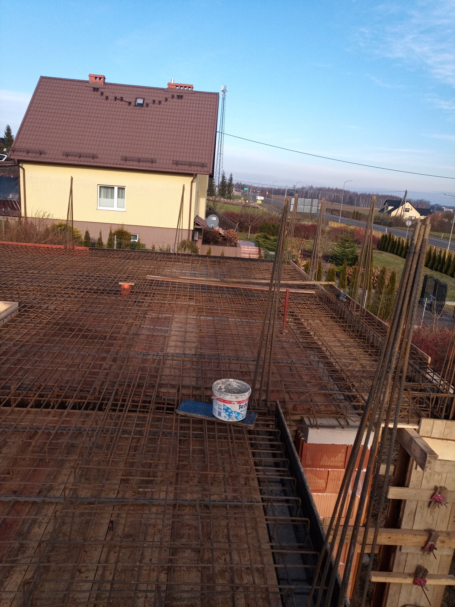 Zbrojenie stropu na budowie domu jednorodzinnego z widoczną siatką stalową, szalunkiem i wiaderkiem z zaprawą murarską na tle żółtej elewacji budynku i błękitnego nieba.