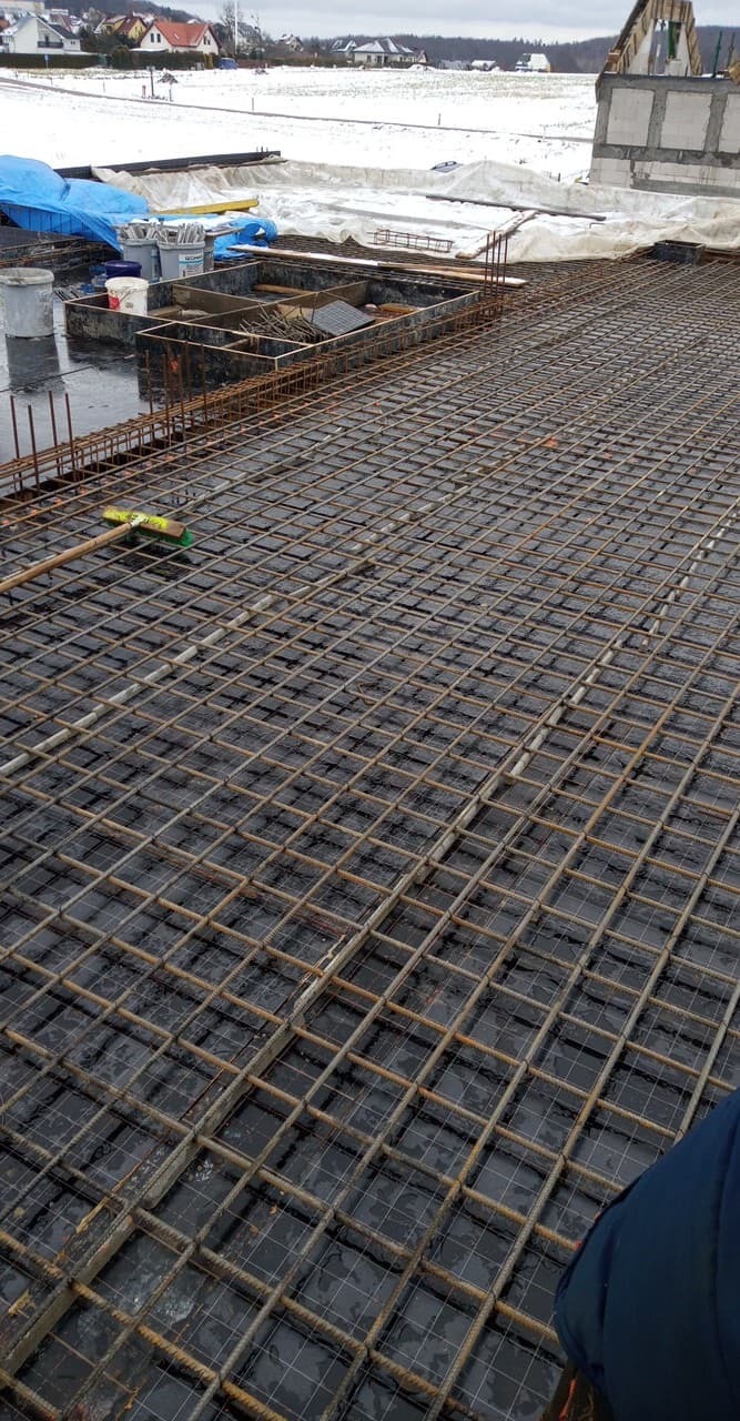 Zbrojenie płyty fundamentowej z widocznymi deskowaniami i przygotowaniami do zalania betonem, w tle zabudowa jednorodzinna w zimowej scenerii.